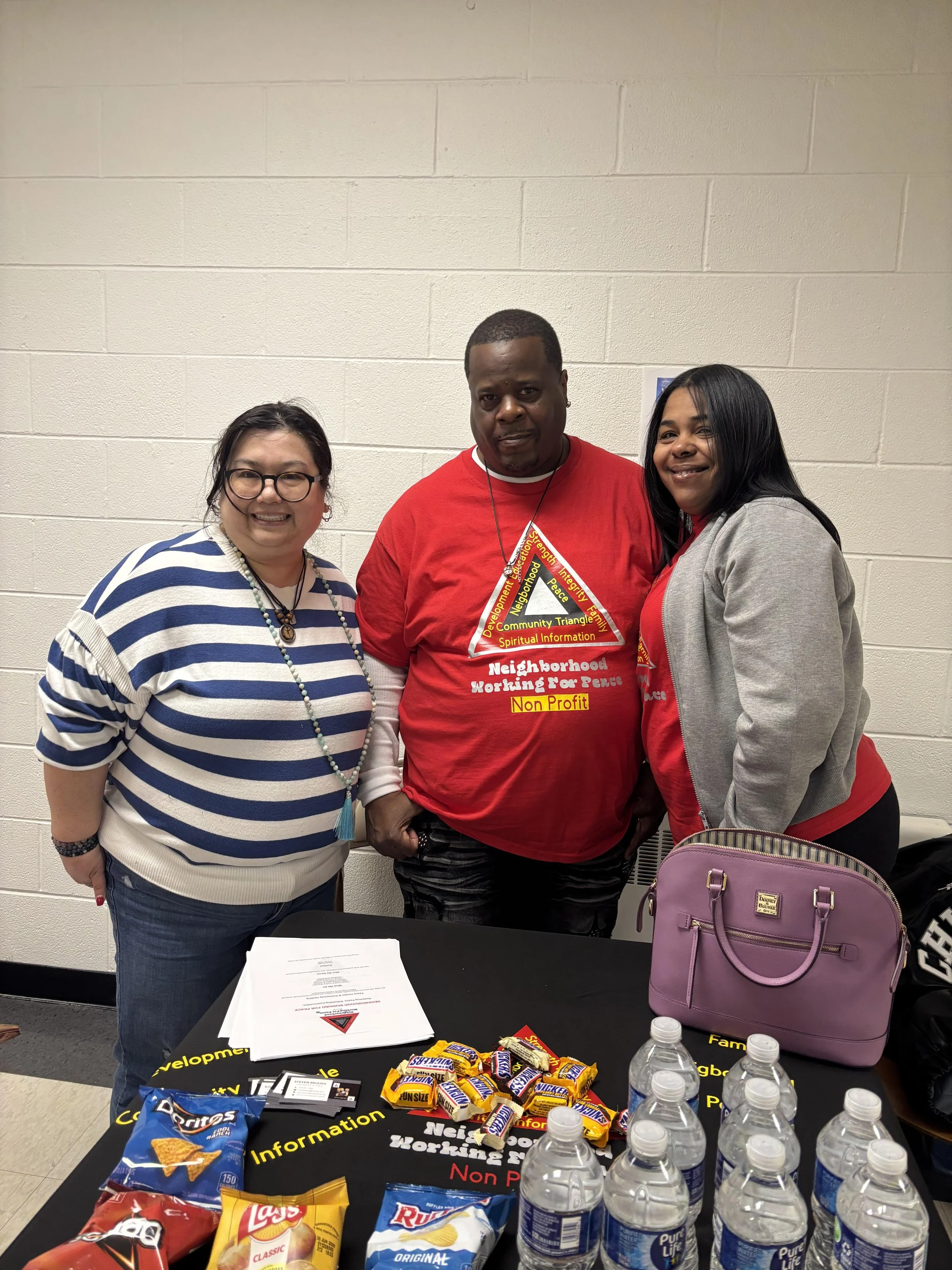 Three people standing behind a table with snacks and bottled water, smiling at the camera, with a plain white brick wall in the background.
