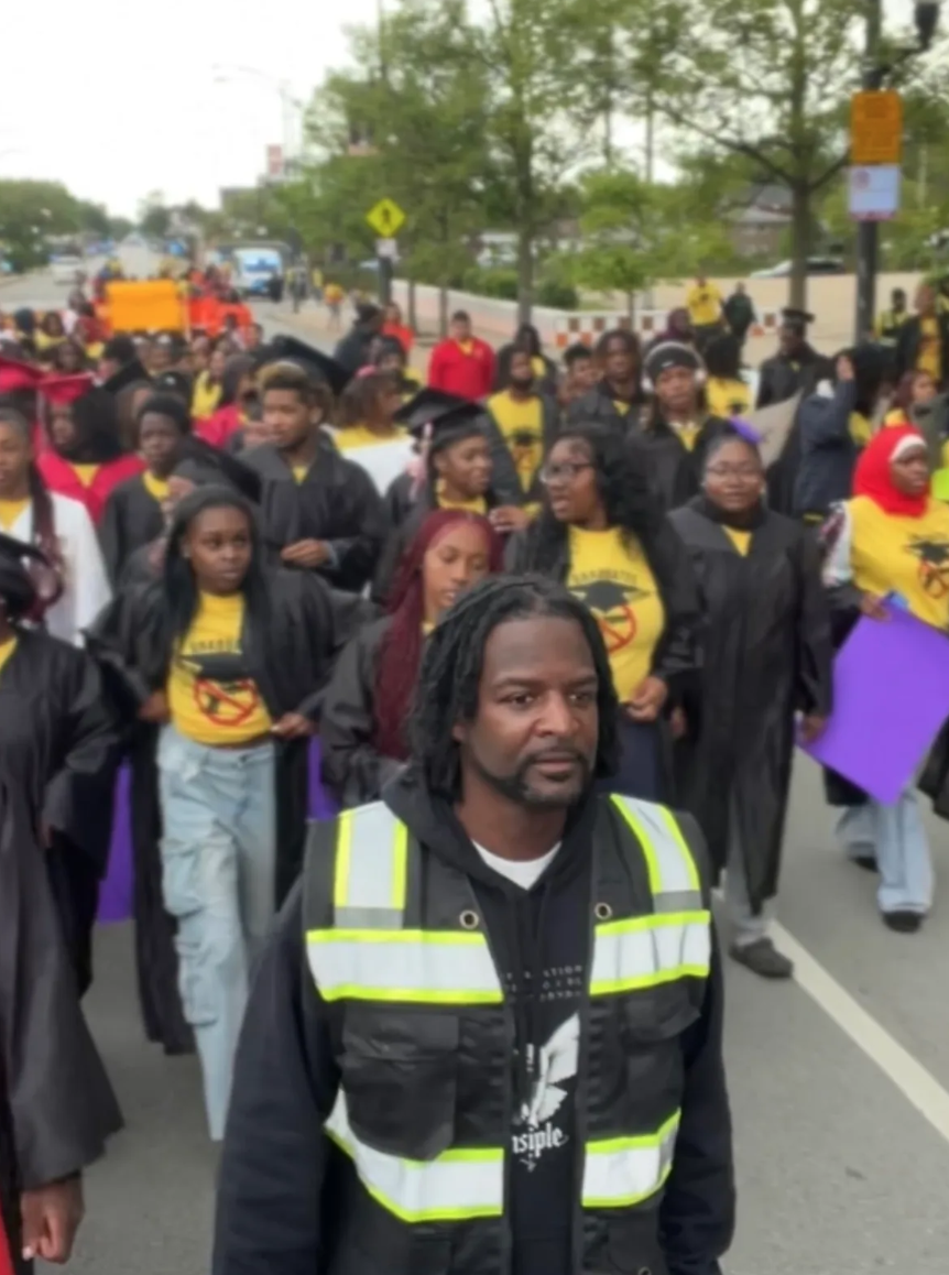 A group of people participating in a protest or march, many wearing black graduation gowns, some holding signs, and a man at the front wearing a black vest with yellow reflective stripes.