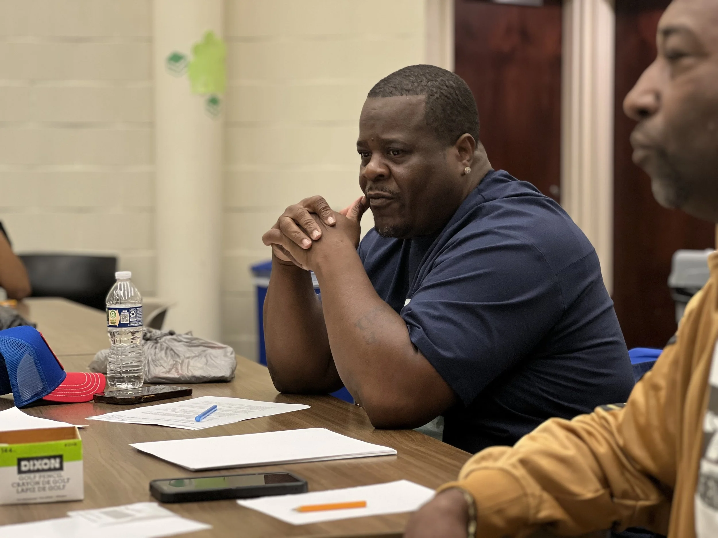 A man with dark skin, short hair, wearing a dark blue shirt, sitting at a table with his hands clasped in front of him, looking contemplative. The table has a water bottle, papers, a pen, and a smartphone. Another person is partially visible on the right side of the image.