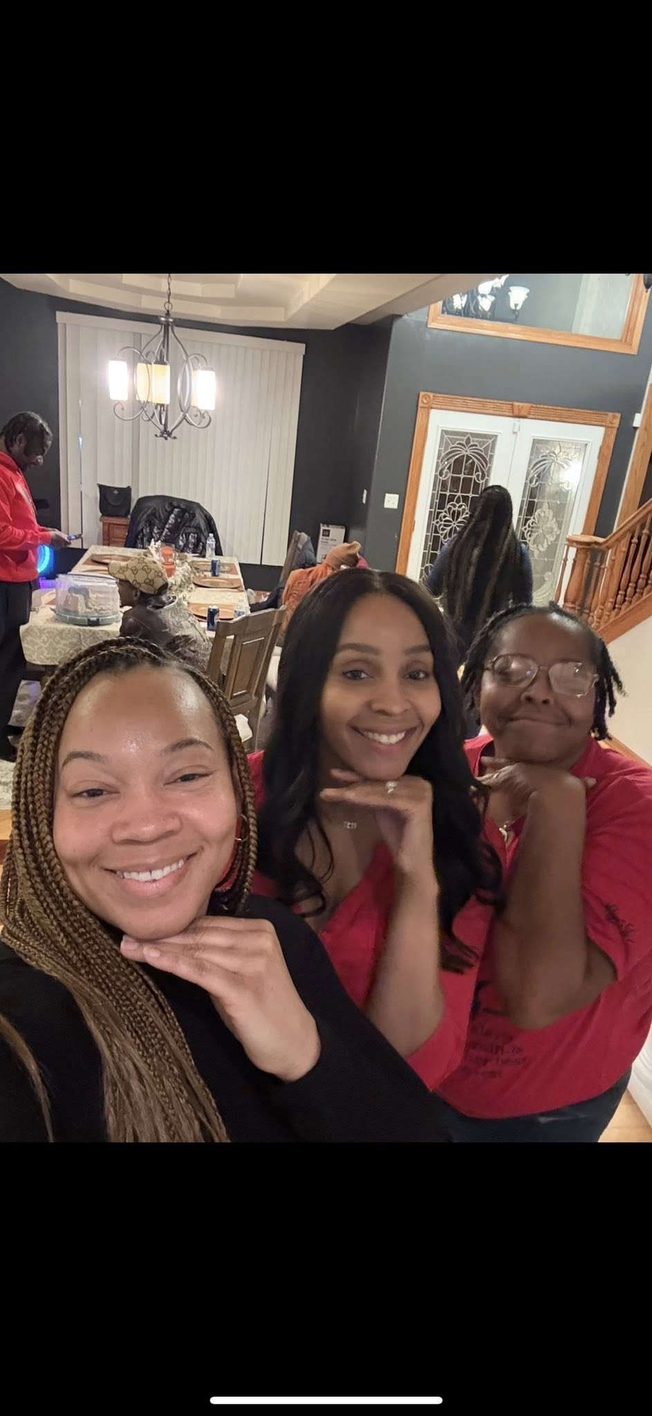 Three women smiling and posing for a photo inside a home, with a dining area and other people in the background.