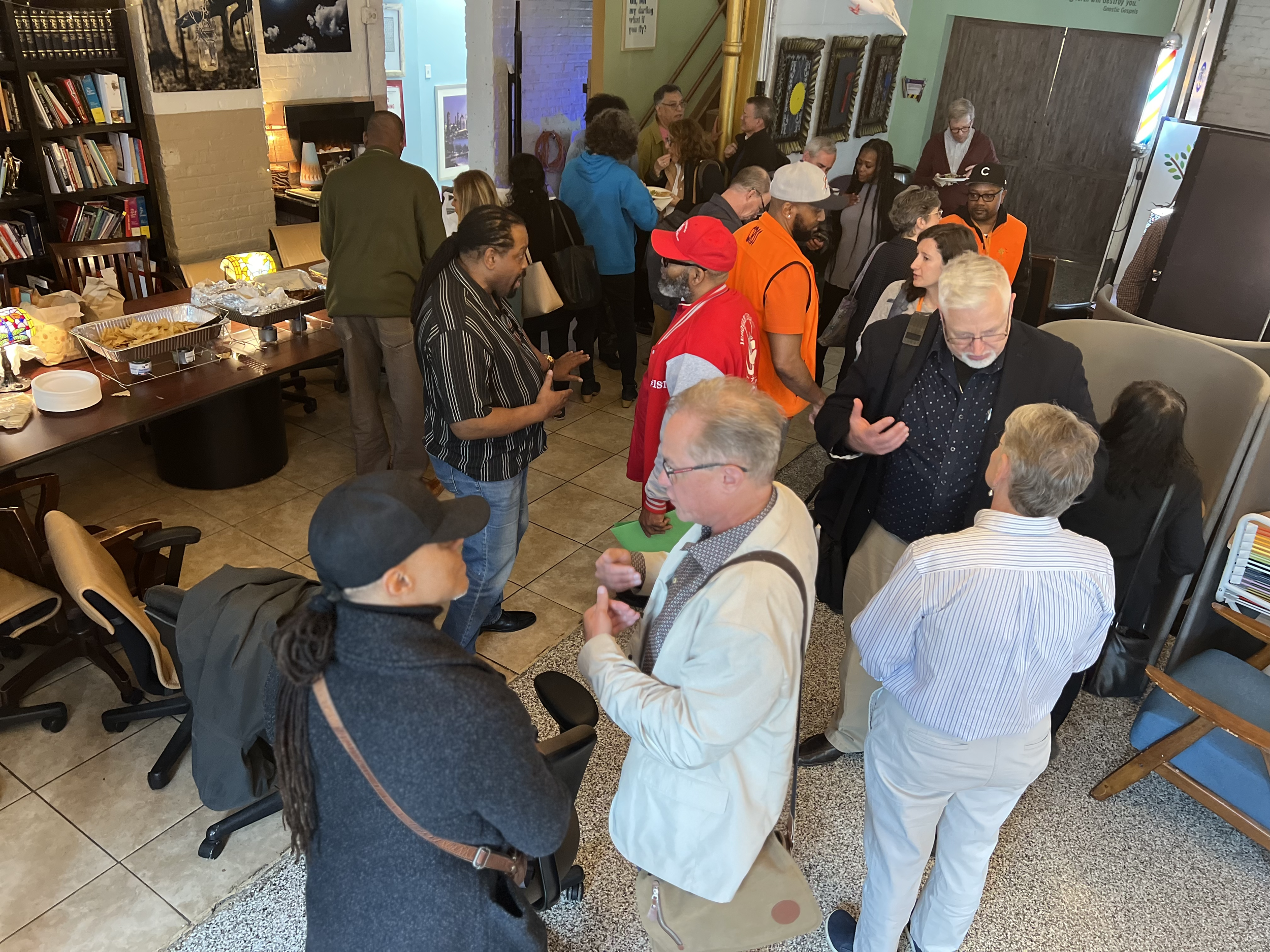 A crowded indoor social gathering with people engaging in conversations. Some are standing near a table with snacks, others are talking in small groups. The setting includes bookshelves, framed pictures, and colorful decor.