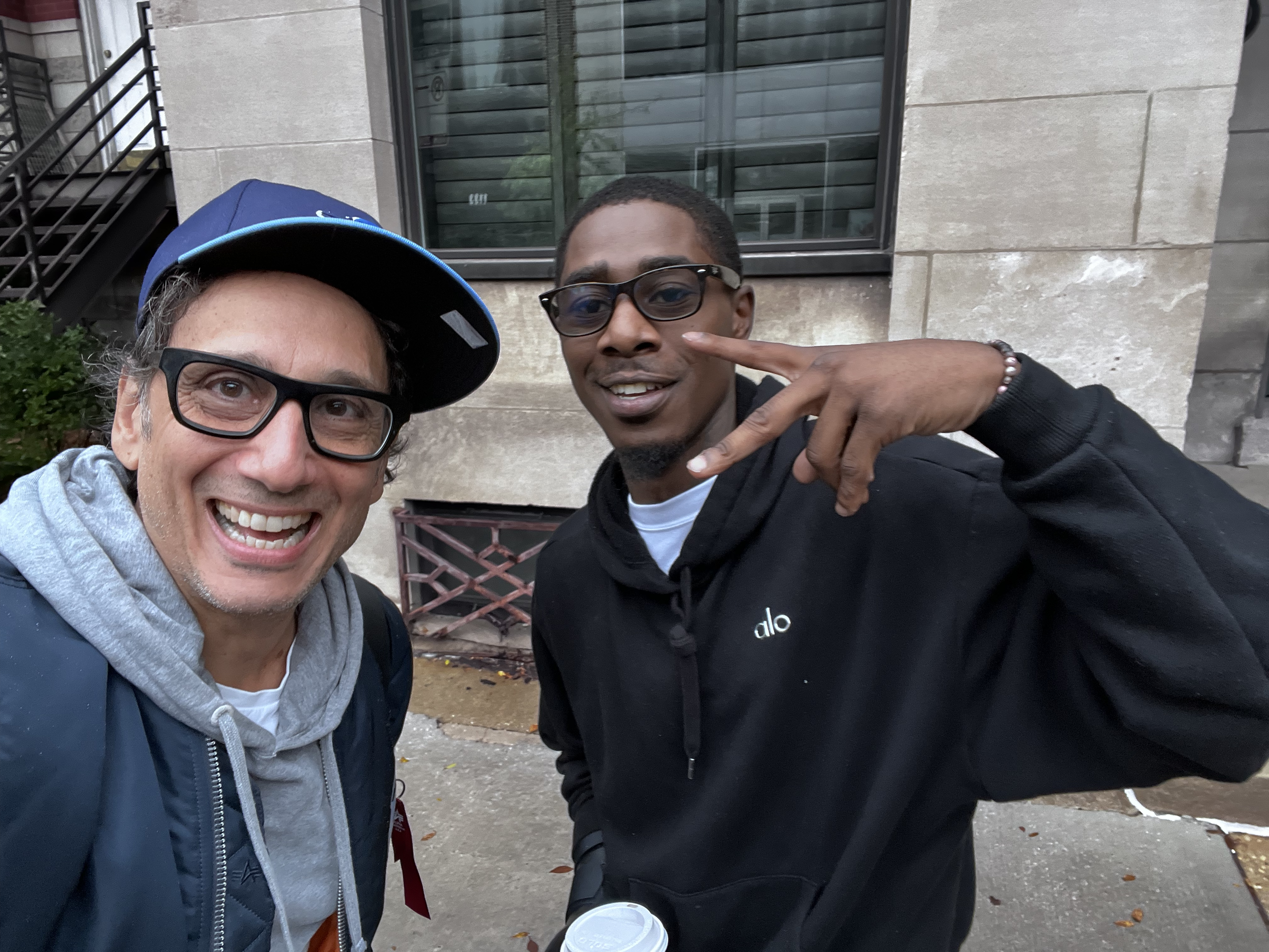 Two men taking a selfie outdoors in front of a building. The man on the left is smiling, wearing glasses, a blue cap sideways, a gray hoodie, and a dark jacket. The man on the right is making a peace sign, wearing glasses, a black hoodie with small white logo, and holding a coffee cup.