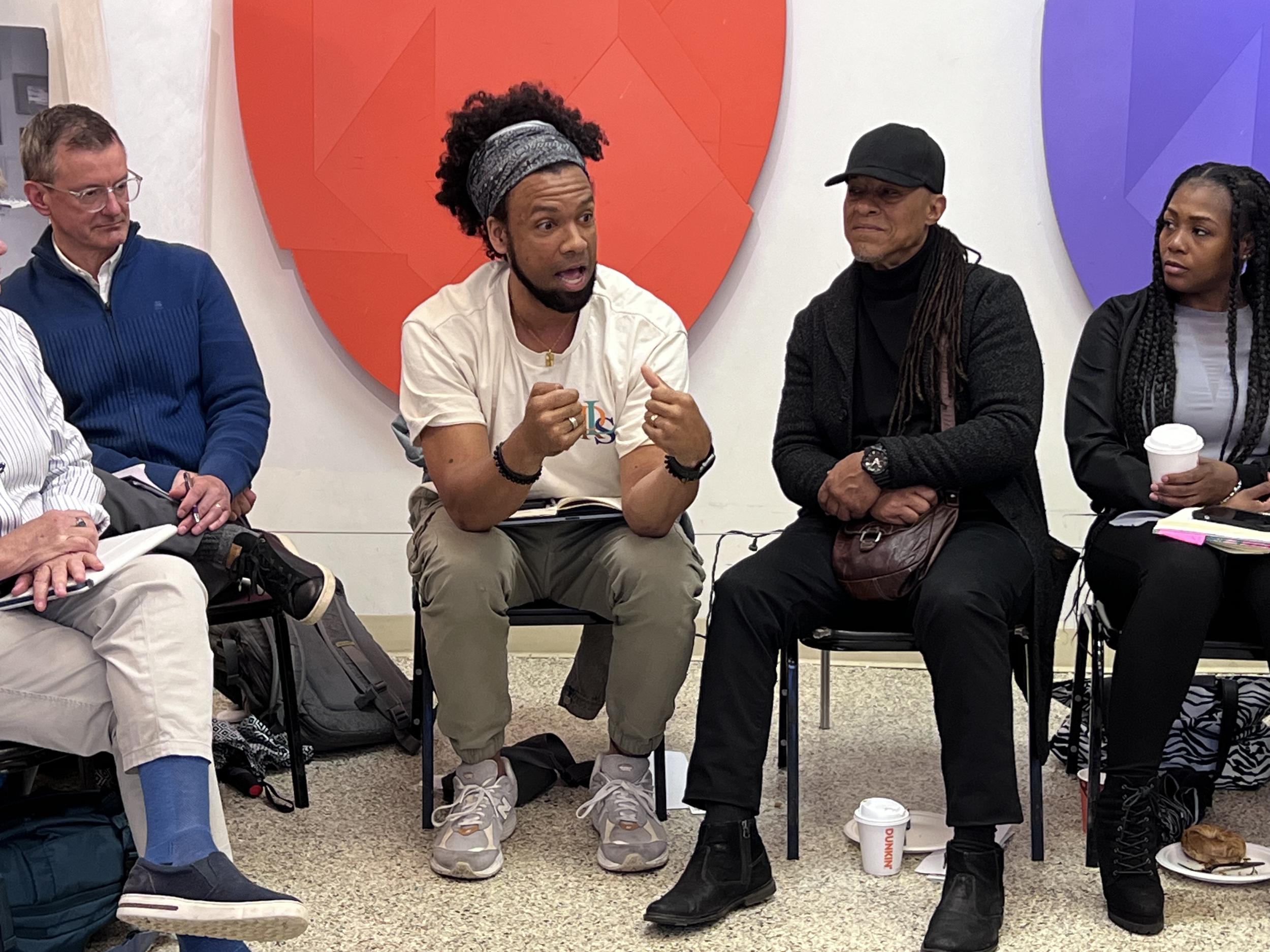 A group of people seated in a circle, participating in a discussion or meeting. The person in the center of the image is speaking, gesturing with hands, wearing a white t-shirt and beige pants. The others are listening, some with notebooks or drinks, in a room with colorful abstract art on the wall.