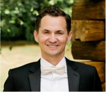 A man in a tuxedo smiling outdoors with a wooden structure and green foliage in the background.