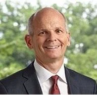 A bald man wearing a dark suit, white shirt, and red tie smiling outdoors with green trees in the background.