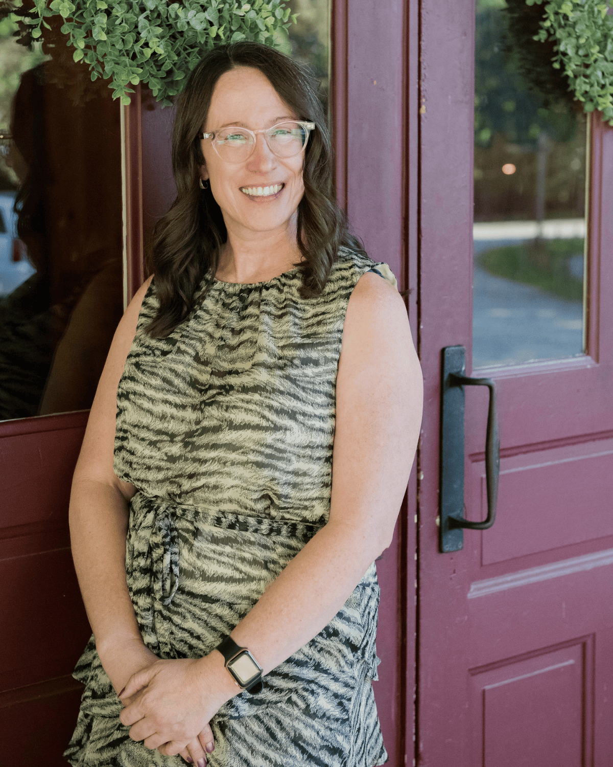 Emily Mendez, mental health copywriter, smiling, wearing a sleeveless animal print dress, standing in front of a maroon door with green hanging plants.