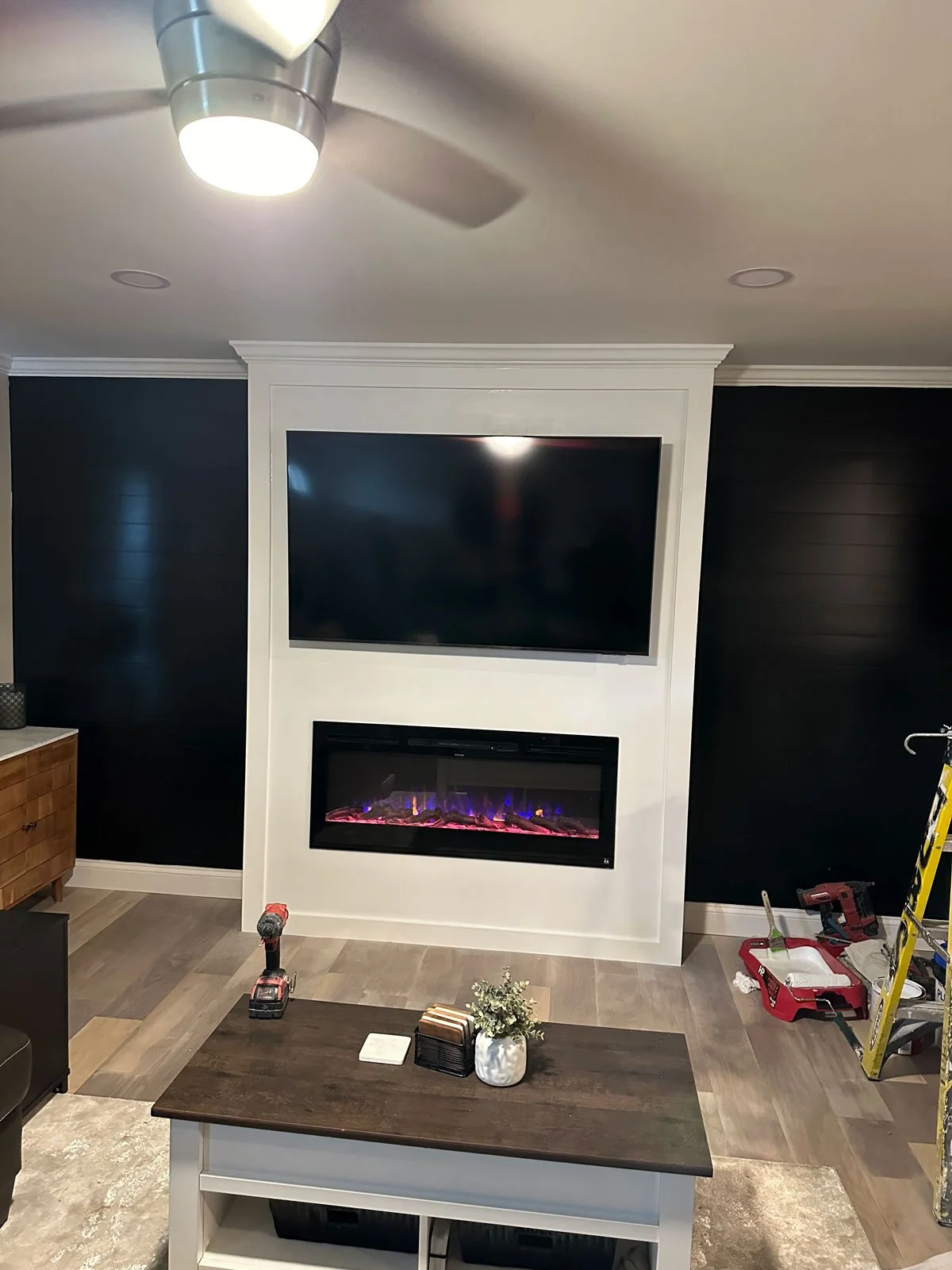 Living room with a ceiling fan, mounted TV, and electric fireplace, with construction tools and equipment visible.