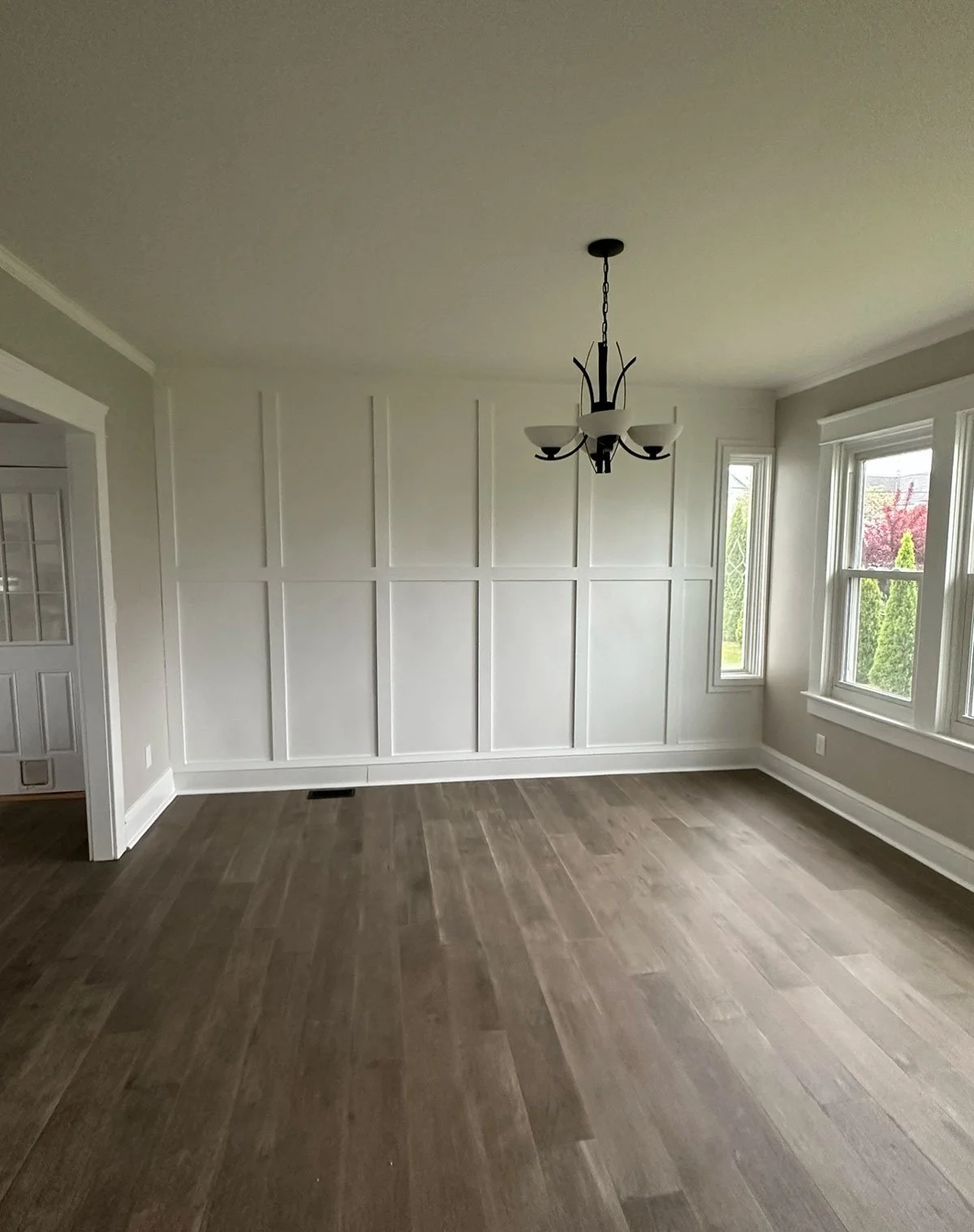 Empty dining room or living space with white walls, large windows, wooden flooring, and a chandelier hanging from the ceiling.