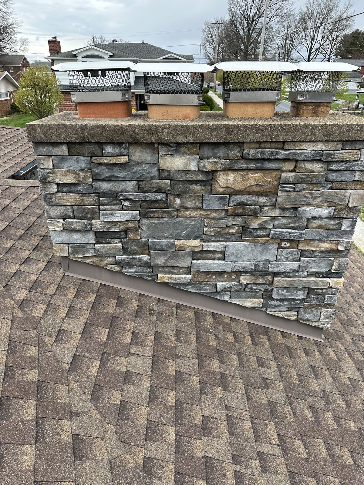 View of a rooftop chimney with a stone exterior, four chimney caps with mesh covers, and a shingled roof, in a suburban neighborhood with houses and trees in the background.