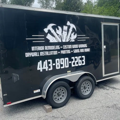 A black trailer with white graphic text and tools, advertising interior remodeling, custom wood working, drywall installation, painting, siding, and more, with a phone number 443-890-2263.