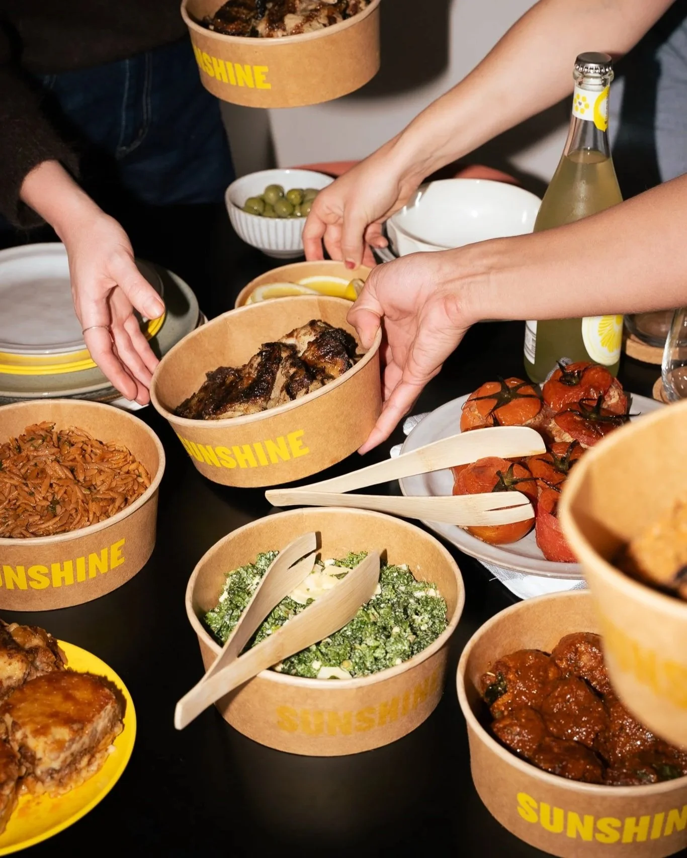 Various dishes of food on a table including bowls of rice, salad, stuffed tomatoes, meat dishes, and a bottle of lemon-flavored beverage, with people reaching for the food.