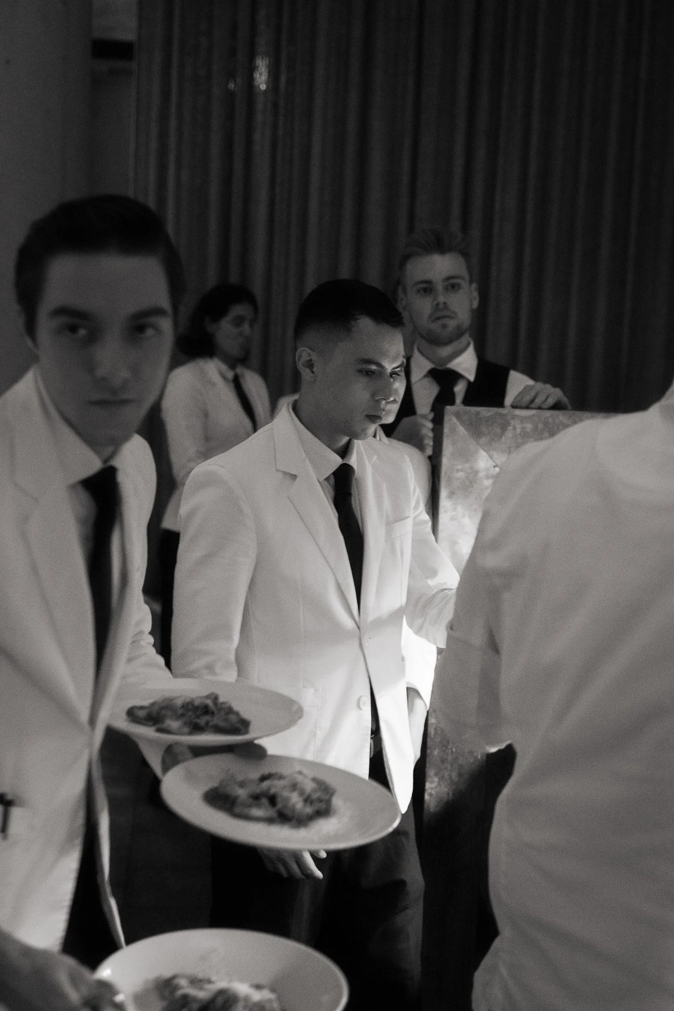 Four young men dressed in white shirts and black ties at a formal event, standing in line while receiving plates of food from a server, with a woman in the background near a curtain.