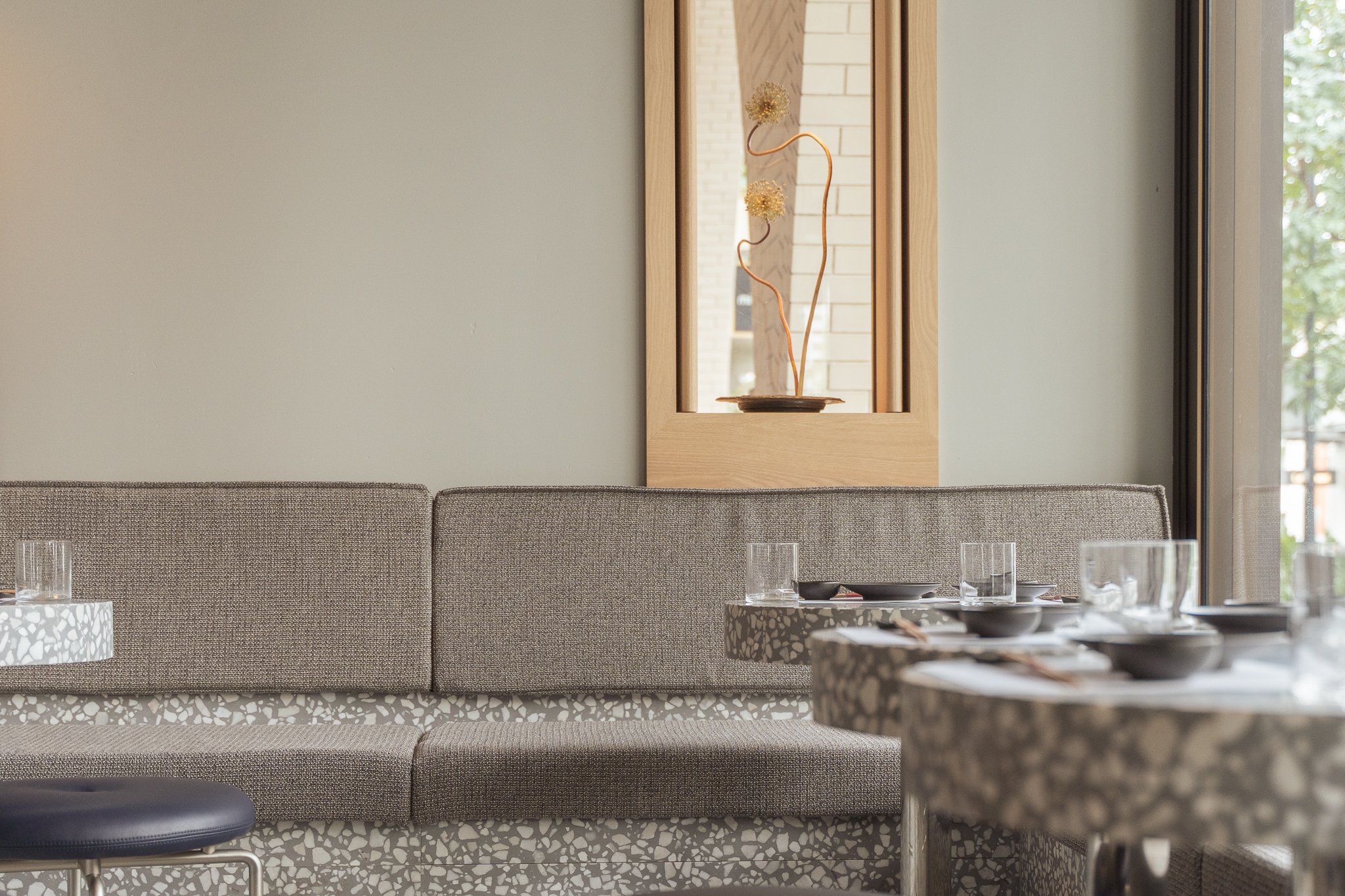 Interior of a dining area with a textured countertop, gray upholstered bench seating, and a glass window. A decorative wall mirror with a wooden frame and art piece inside can be seen in the background.