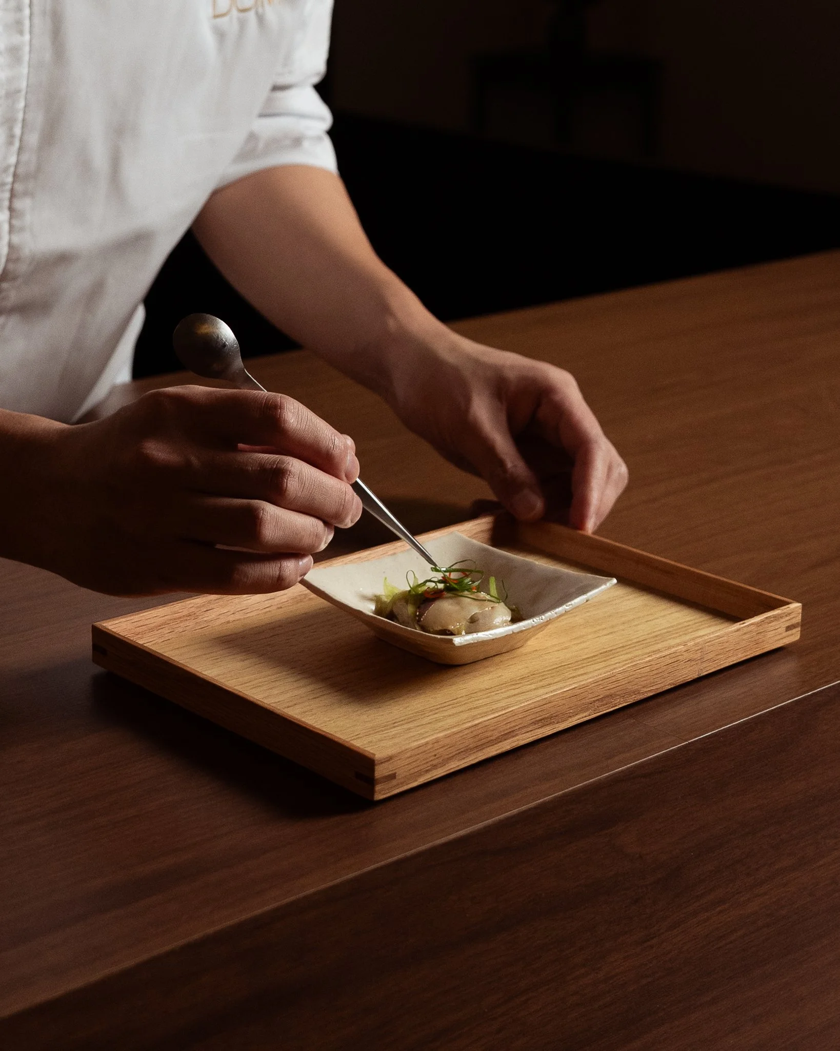 A person in a white shirt is using a spoon to plate food onto a small rectangular dish, which is on a wooden tray on a dark wooden table.