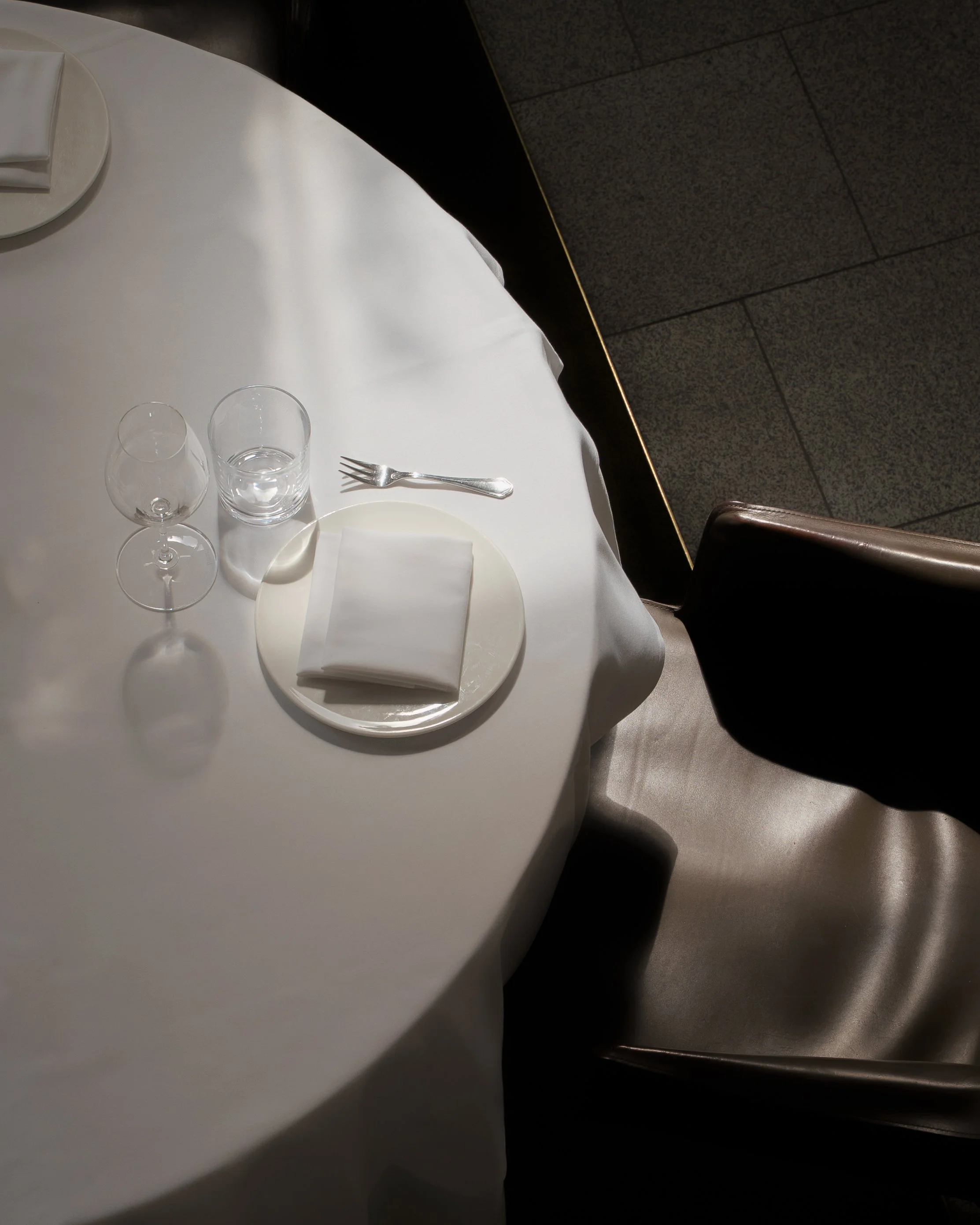 Table with a white tablecloth set with two wine glasses, one water glass, a fork, a plate, a folded napkin, and a small plate with folded white napkins. Part of a chair is visible at the bottom right corner.
