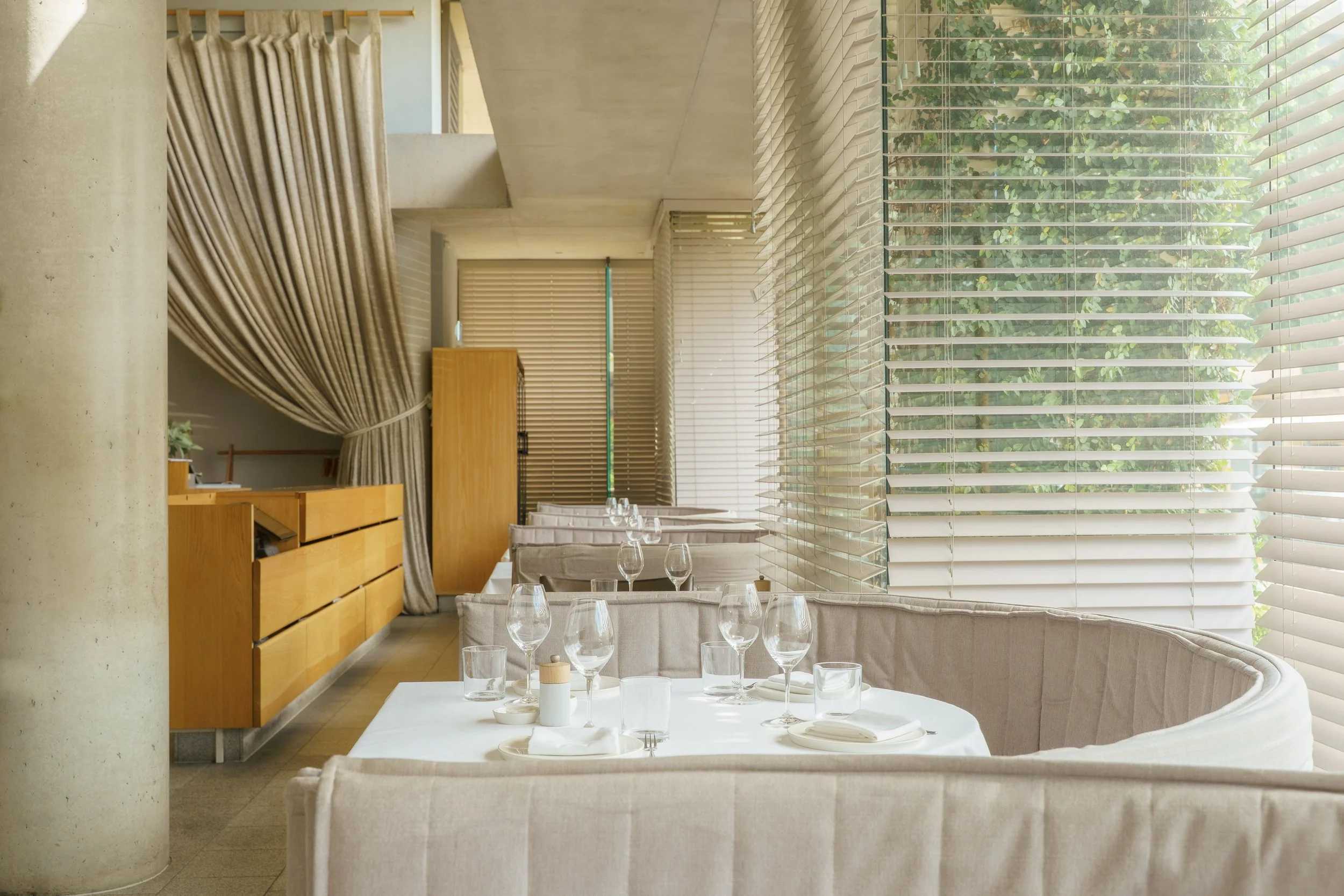 Interior of a modern restaurant with large windows, curved beige booth seating, tables set with glassware, and wooden cabinetry, illuminated by natural light from outside.