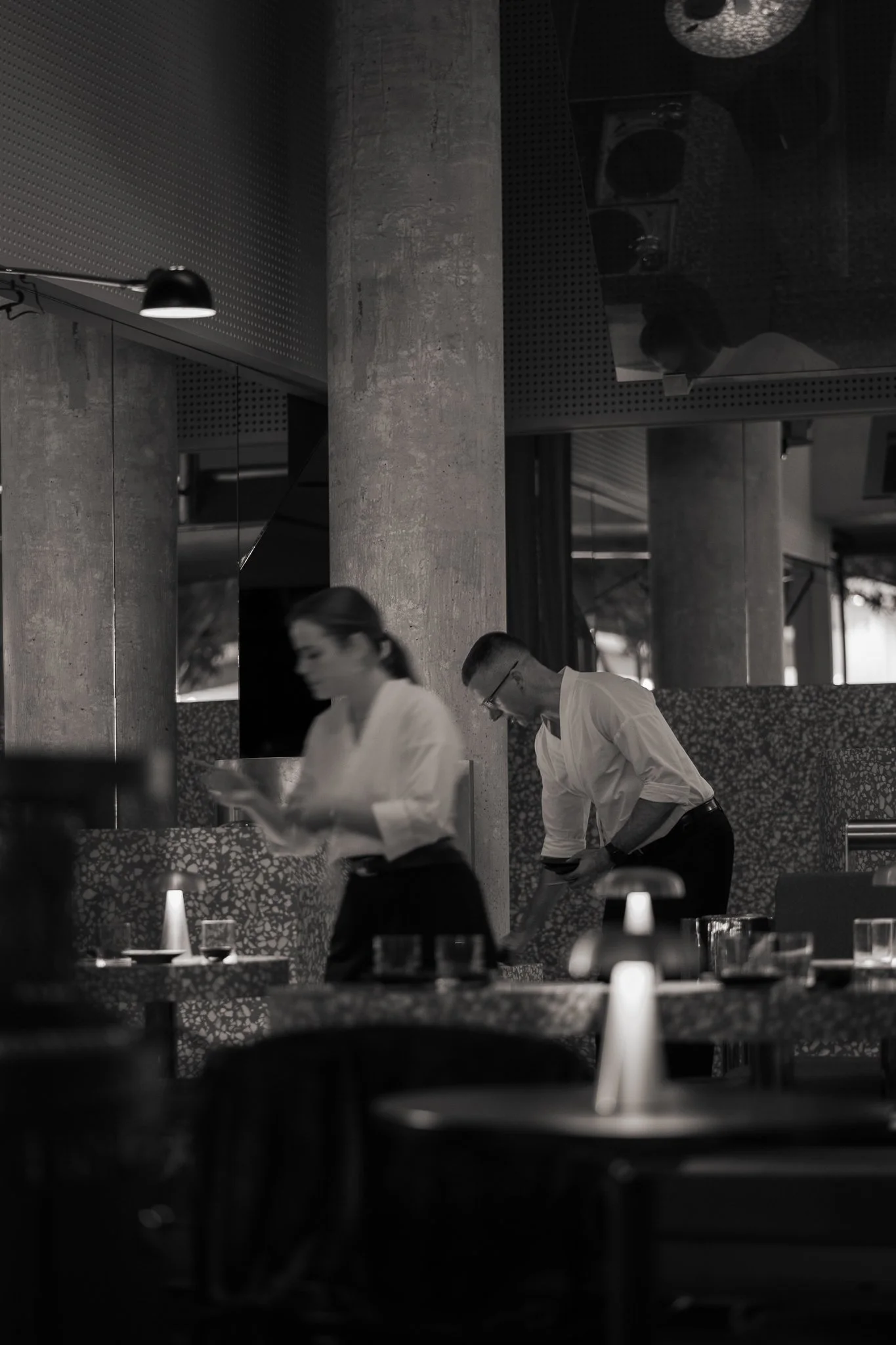 Black and white photo of a modern restaurant interior with several people, including a woman and two men in business attire, engaging in activities like reading or working. The setting includes concrete columns, patterned wallpaper, and dining tables