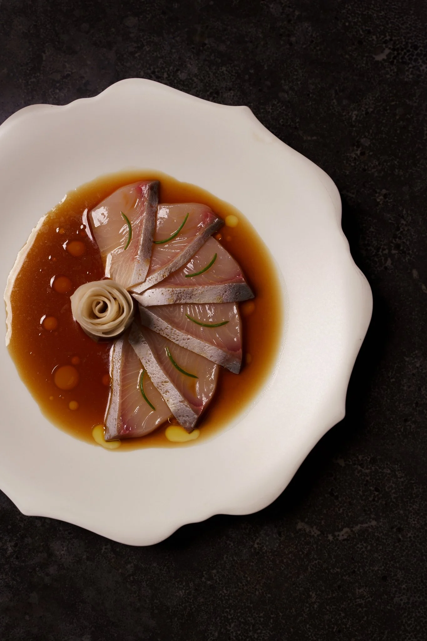 Slices of raw fish, garnished with herbs and served in a soy-based sauce on a white, scalloped-edge plate against a dark countertop.