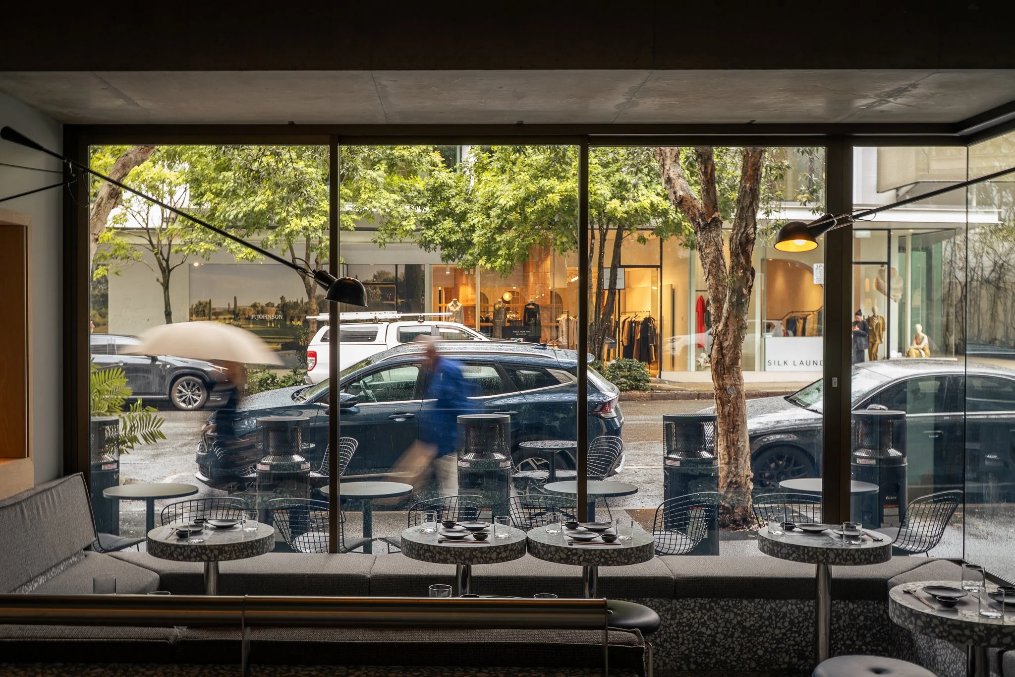 Interior of a modern café with large floor-to-ceiling windows showing trees, parked cars, and a busy street outside. The café has small round tables with black wire chairs, and a gray upholstered bench along the window. Dim lighting fixtures hang from the ceiling.