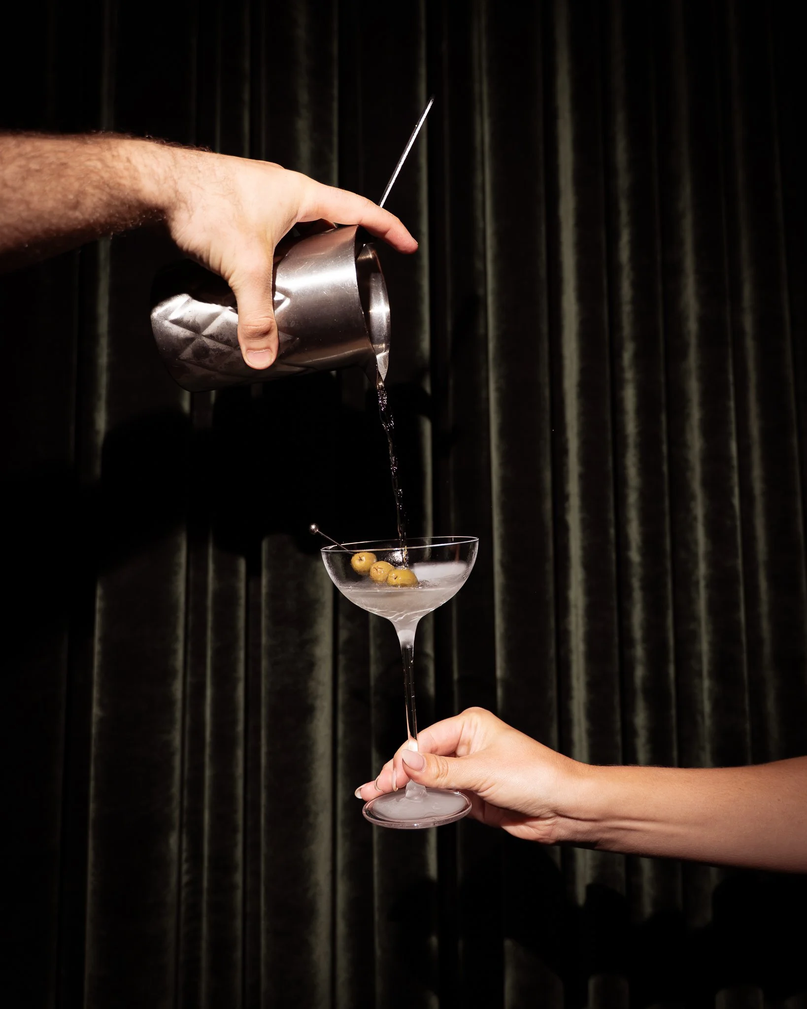 A hand holding a cocktail glass with green olives while pouring liquid from a cocktail shaker, with a dark background and a curtain.
