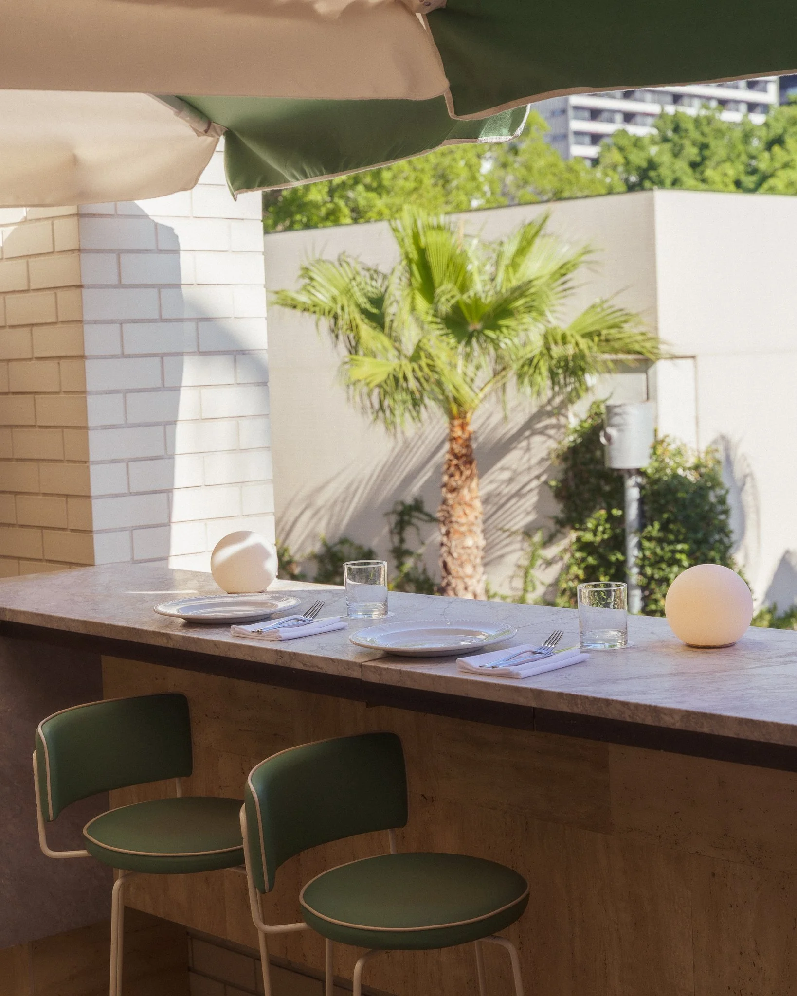 Empty restaurant table set with plates, glasses, and silverware, with green chairs around it. Outside, there is a palm tree and modern buildings in the background.