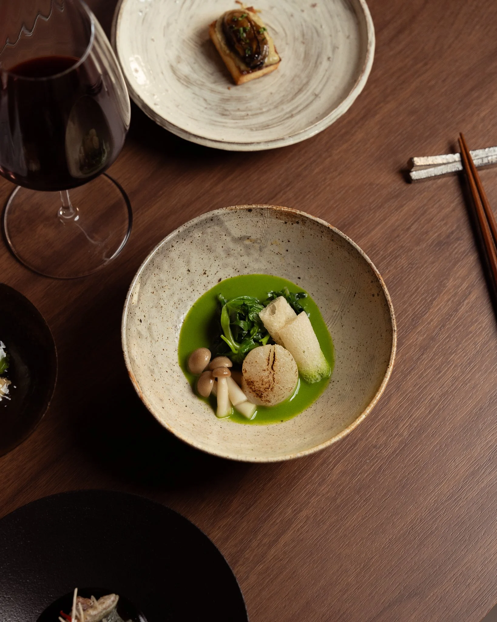A dinner table with a glass of red wine, a plate with a small appetizer, and a bowl of green soup with vegetables and seafood.