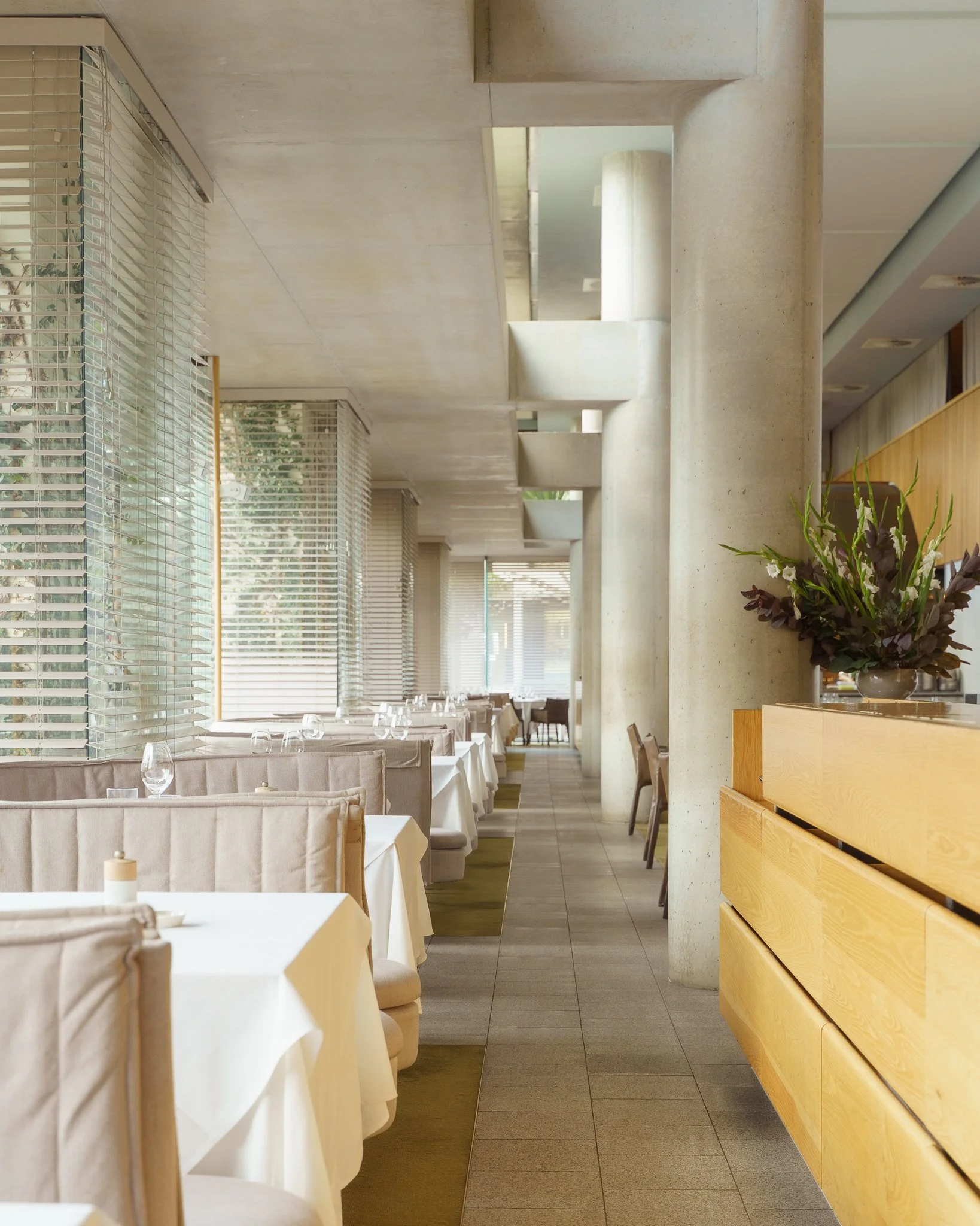 Elegant restaurant interior with white tablecloths, chairs, and large windows with blinds. Modern decor with concrete columns, wooden accents, and a flower arrangement in the foreground.
