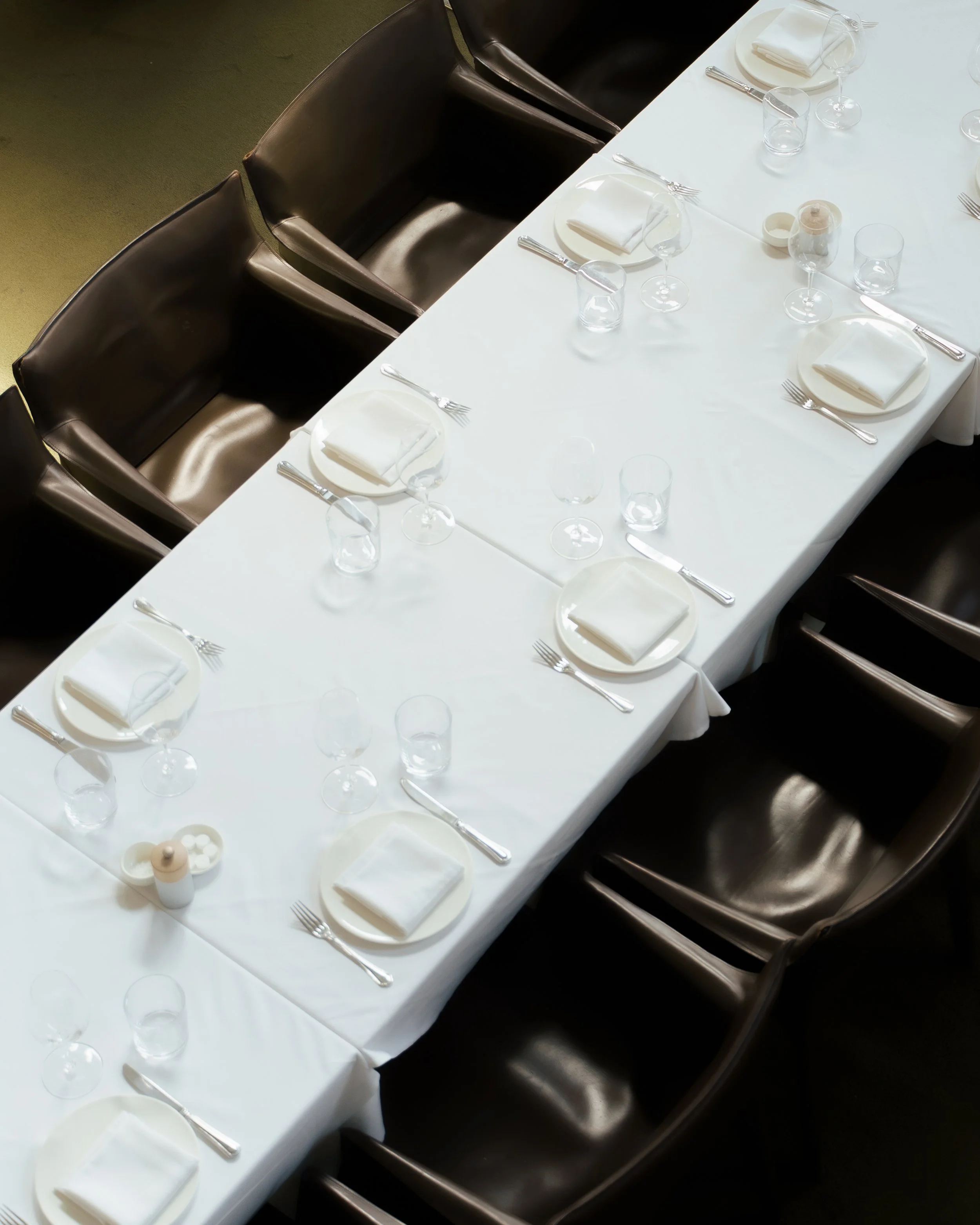 A formal dining table set with white tablecloth, white plates with folded white napkins, silver cutlery, empty wine glasses, water glasses, and small condiment containers, with dark leather chairs around it.