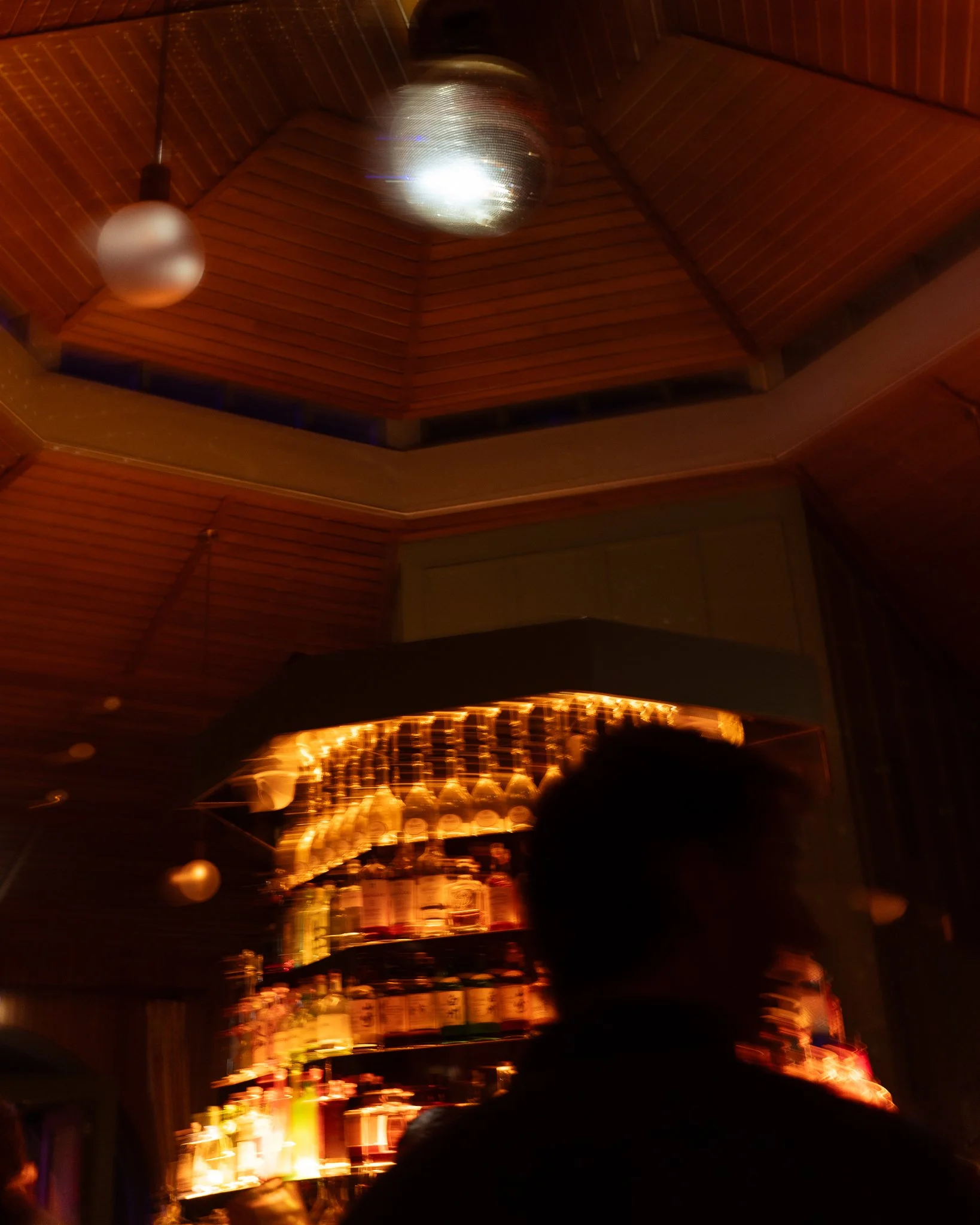 Dimly lit bar or lounge with wooden ceiling and walls, illuminated by warm yellow and orange string lights, with bottles and glasses on shelves in the background.
