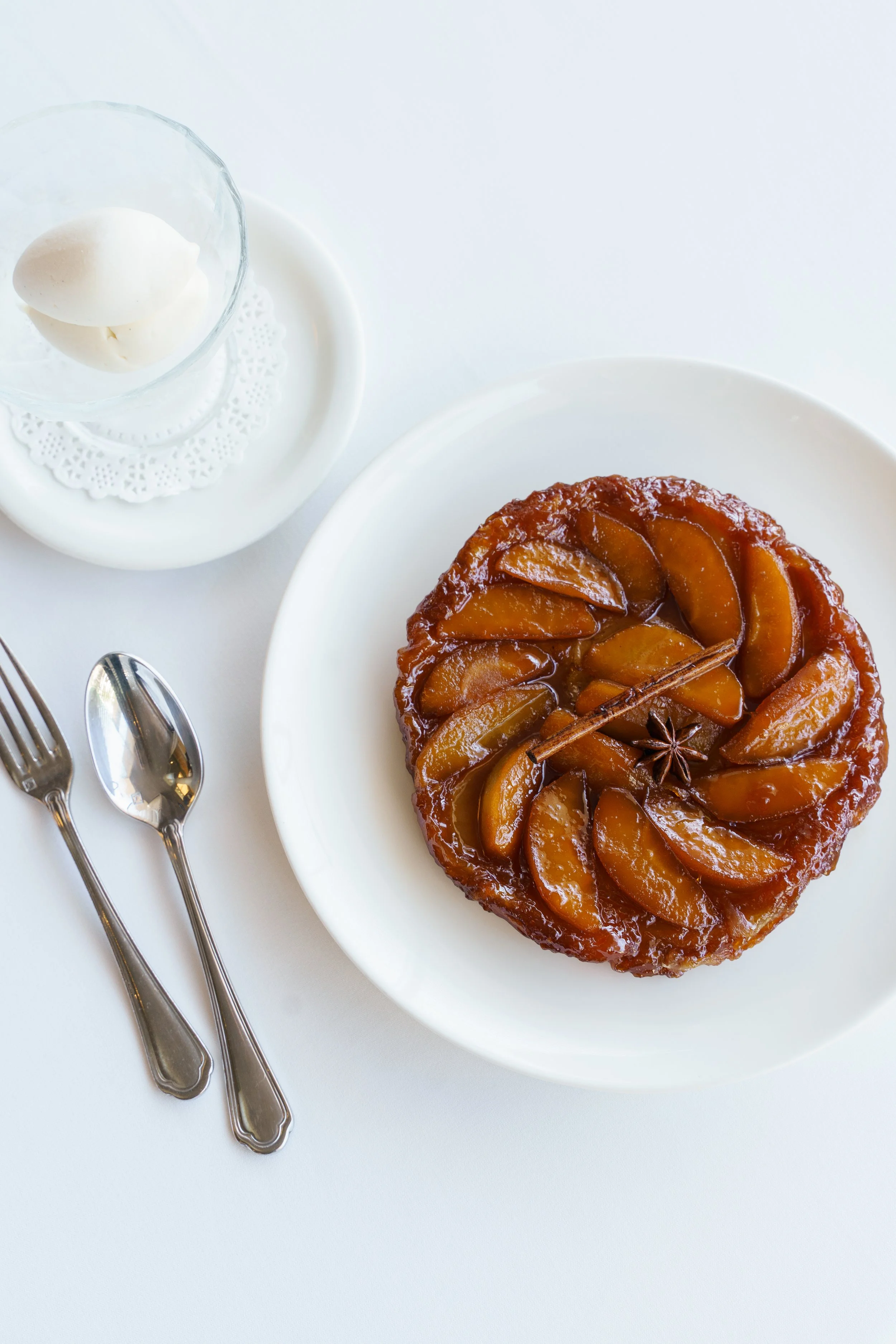 A plate of tart tatin with caramel glaze garnished with star anise, a glass bowl of whipped cream on a plate with a doily, and silverware on a white tablecloth.