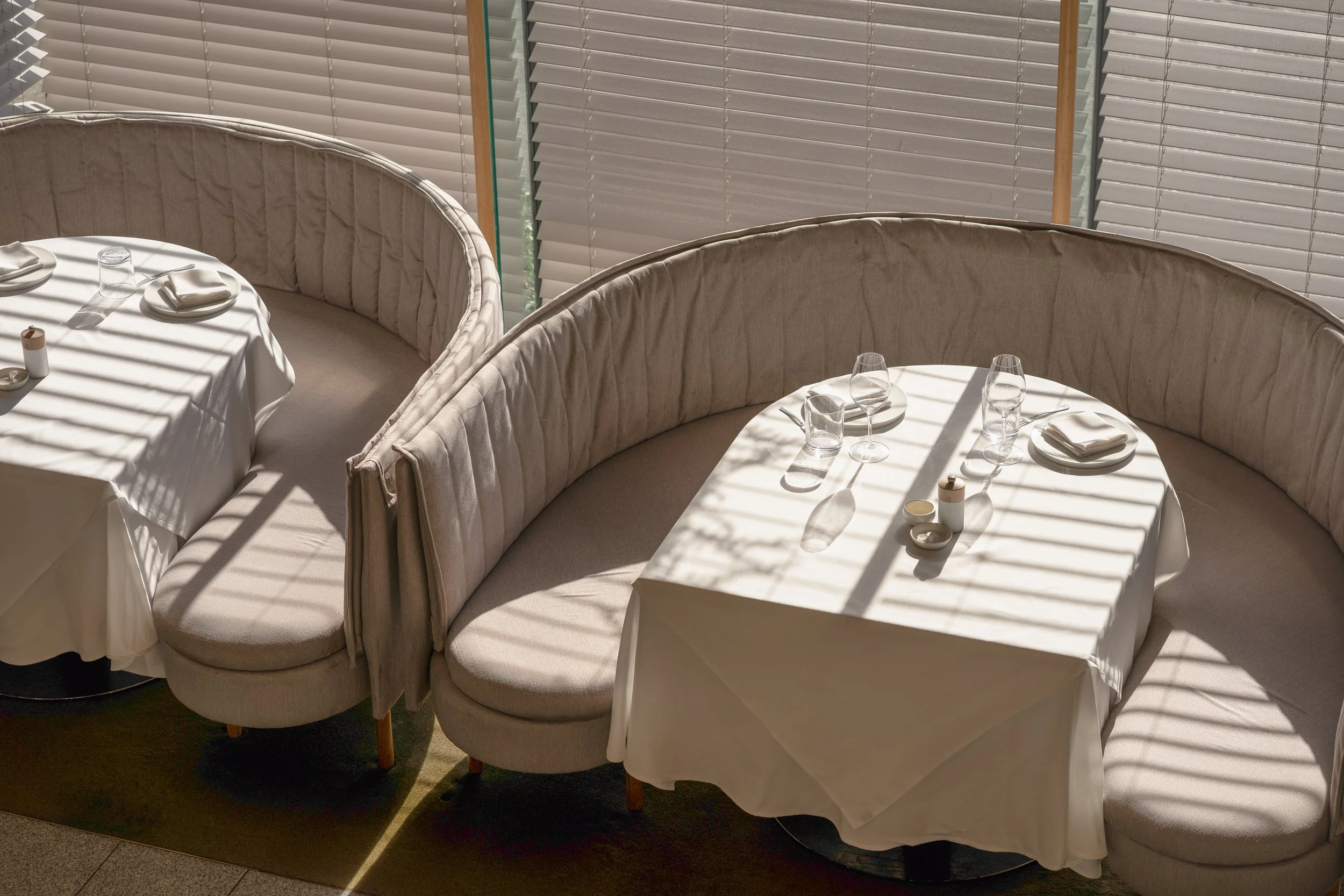 Round dining tables covered with white tablecloths, set with glasses, plates, napkins, and small condiment jars, illuminated by sunlight through window blinds.