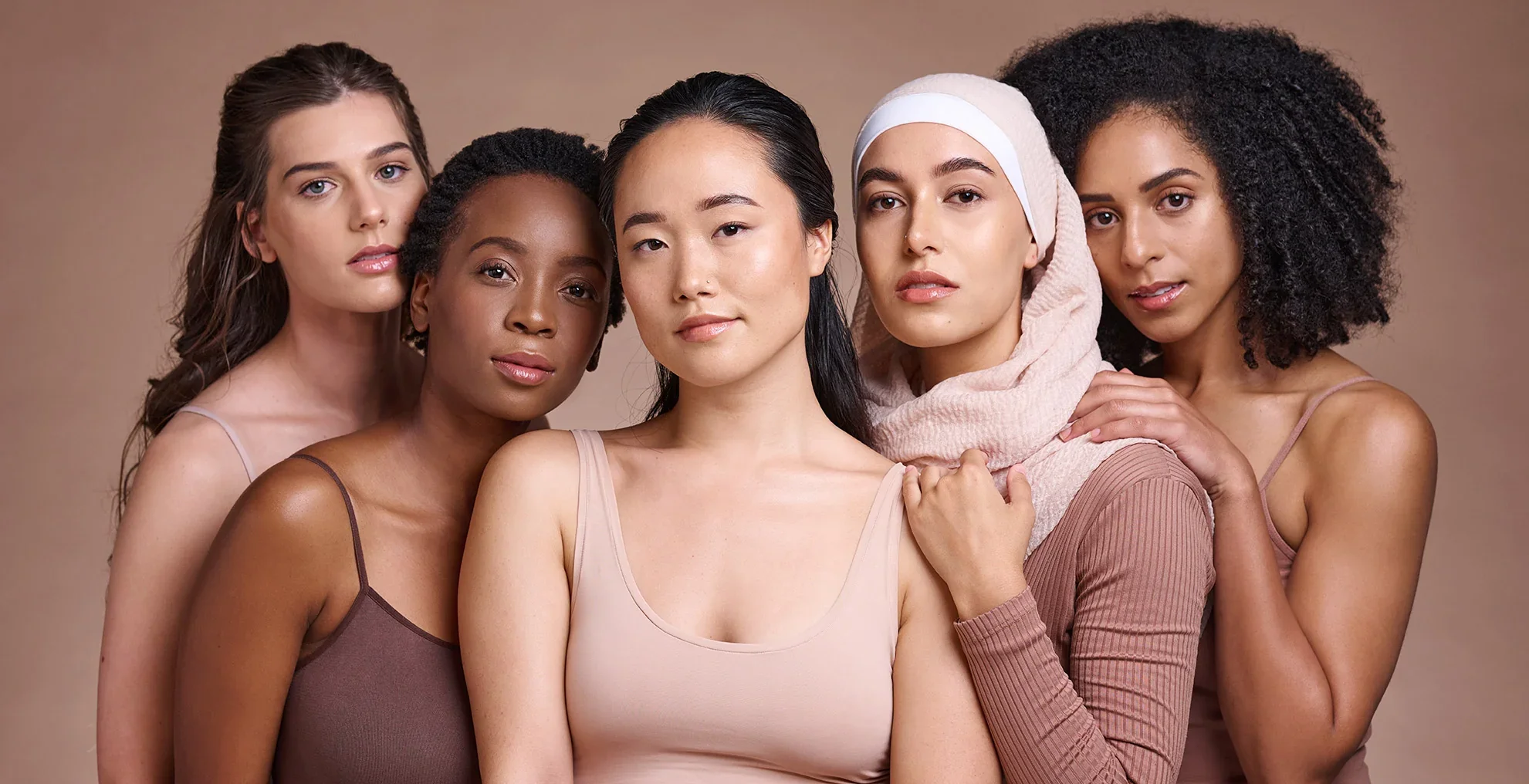 A diverse group of five women standing close together against a neutral background, looking at the camera.