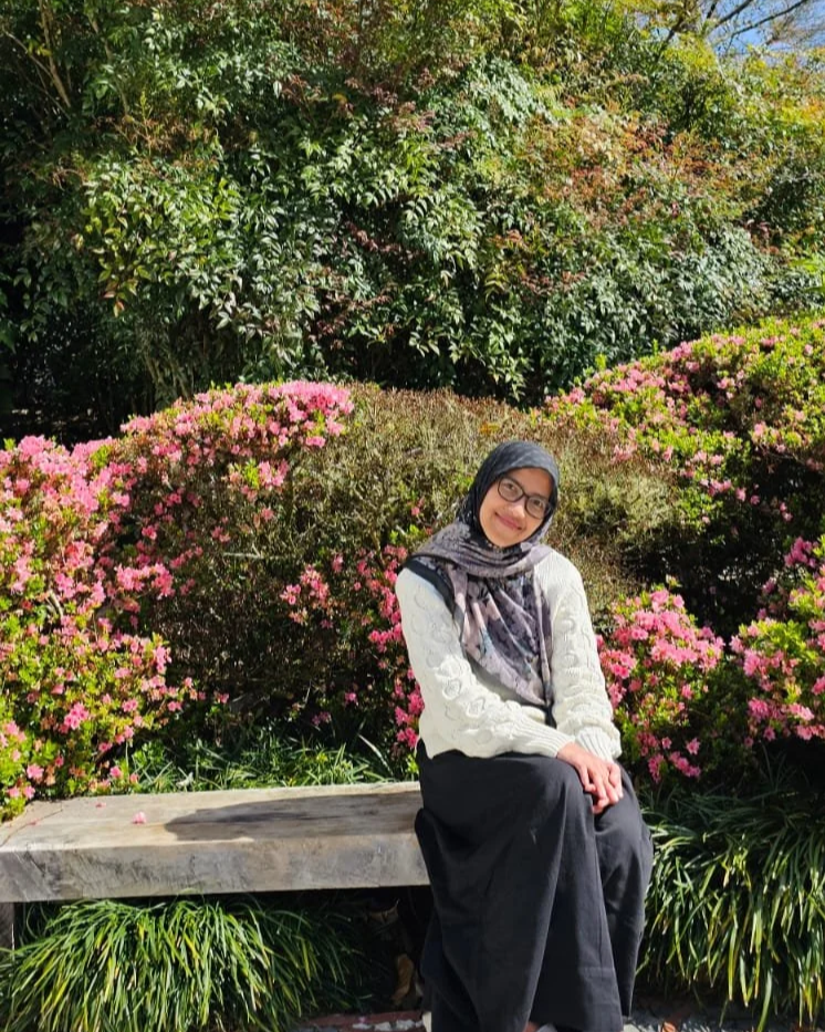 A woman with glasses wearing a black and gray headscarf and a white sweater, sitting on a wooden bench in a garden with pink flowers and green bushes.