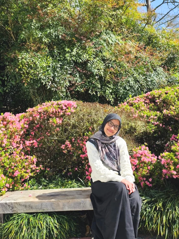 A woman wearing glasses and a hijab sitting on a wooden bench in a garden with pink azaleas and green shrubs, under a partly cloudy sky.