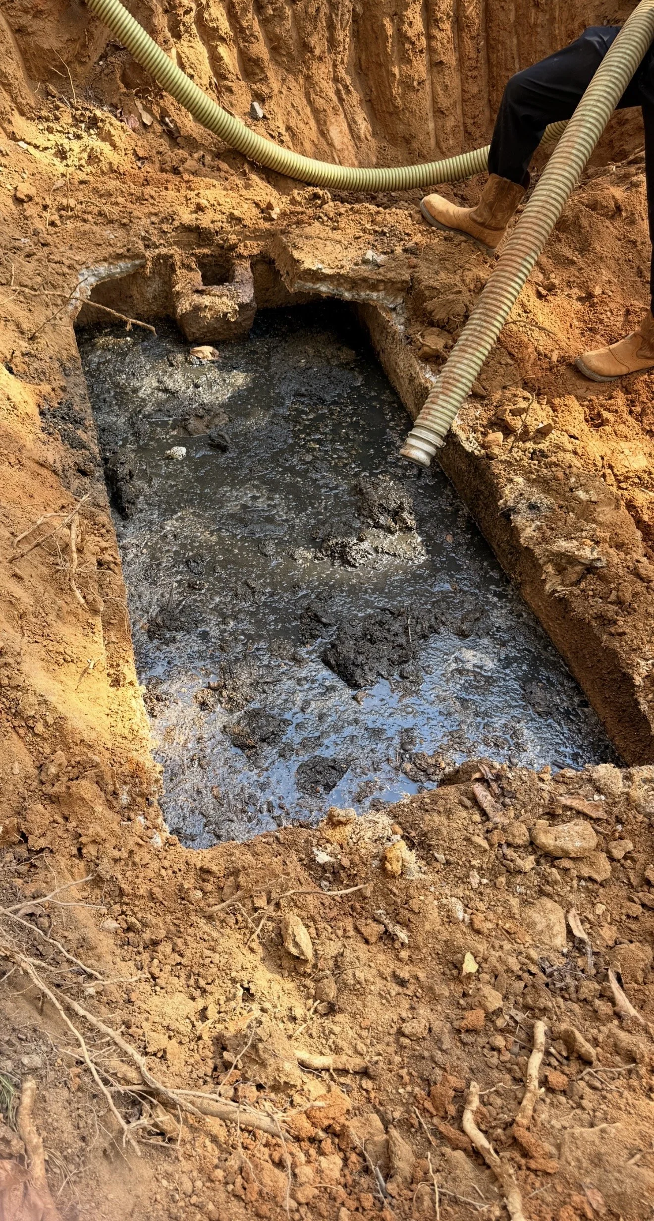 A worker in boots is installing or repairing underground piping with large flexible hoses, with a trench filled with muddy water and surrounding soil.