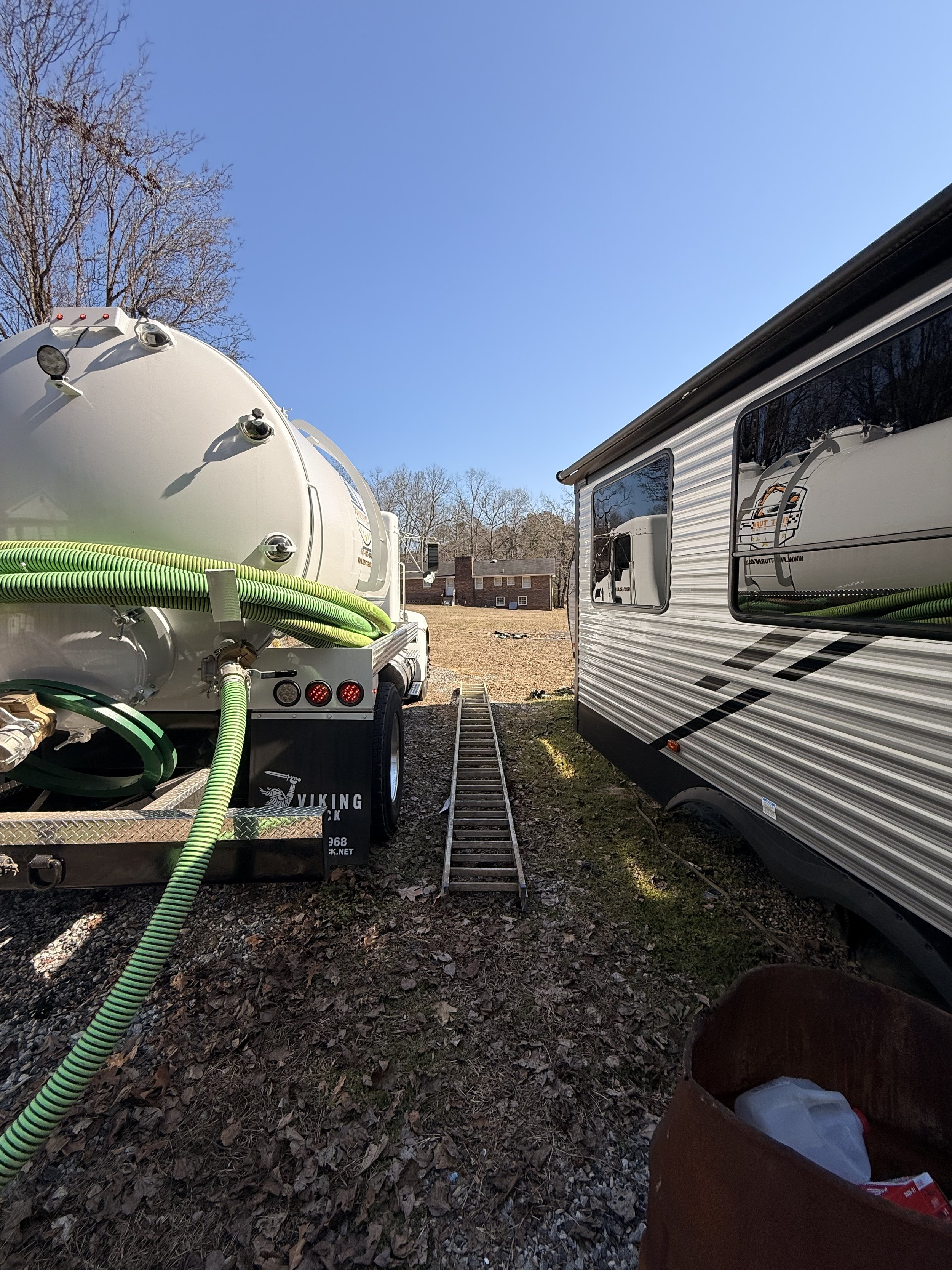 First Turn Septic truck next to an RV parked on a grassy area with a small ladder between them and a clear blue sky overhead.