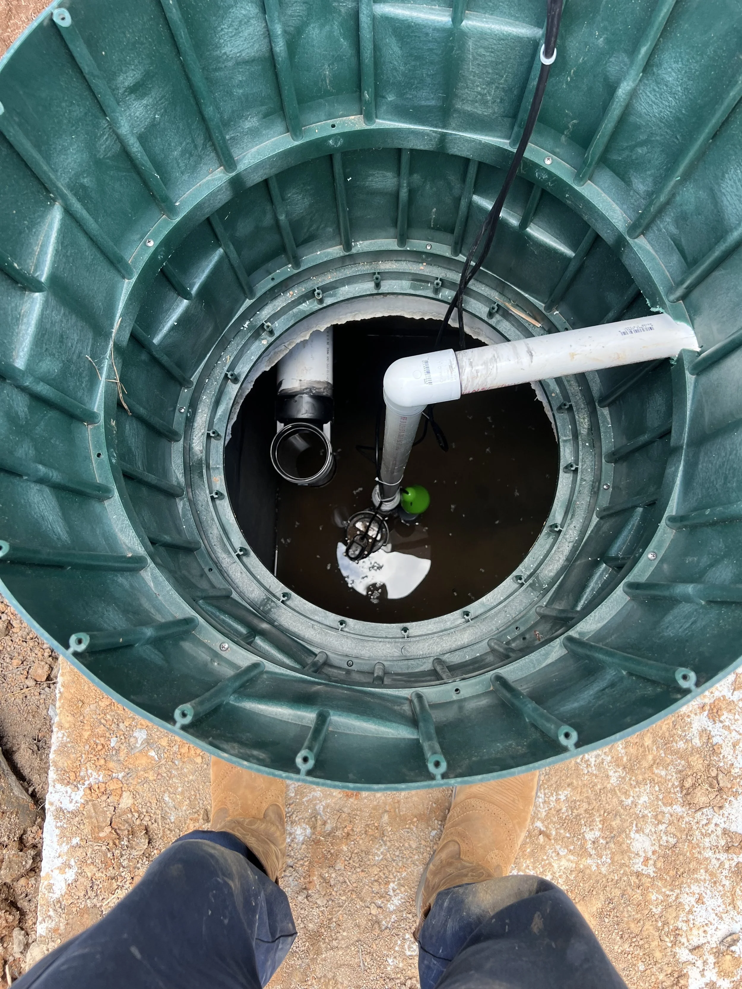 View looking down into a well or a large tank with water at the bottom, containing pipes and inflatables, with a person standing at the edge wearing brown boots and dark jeans.