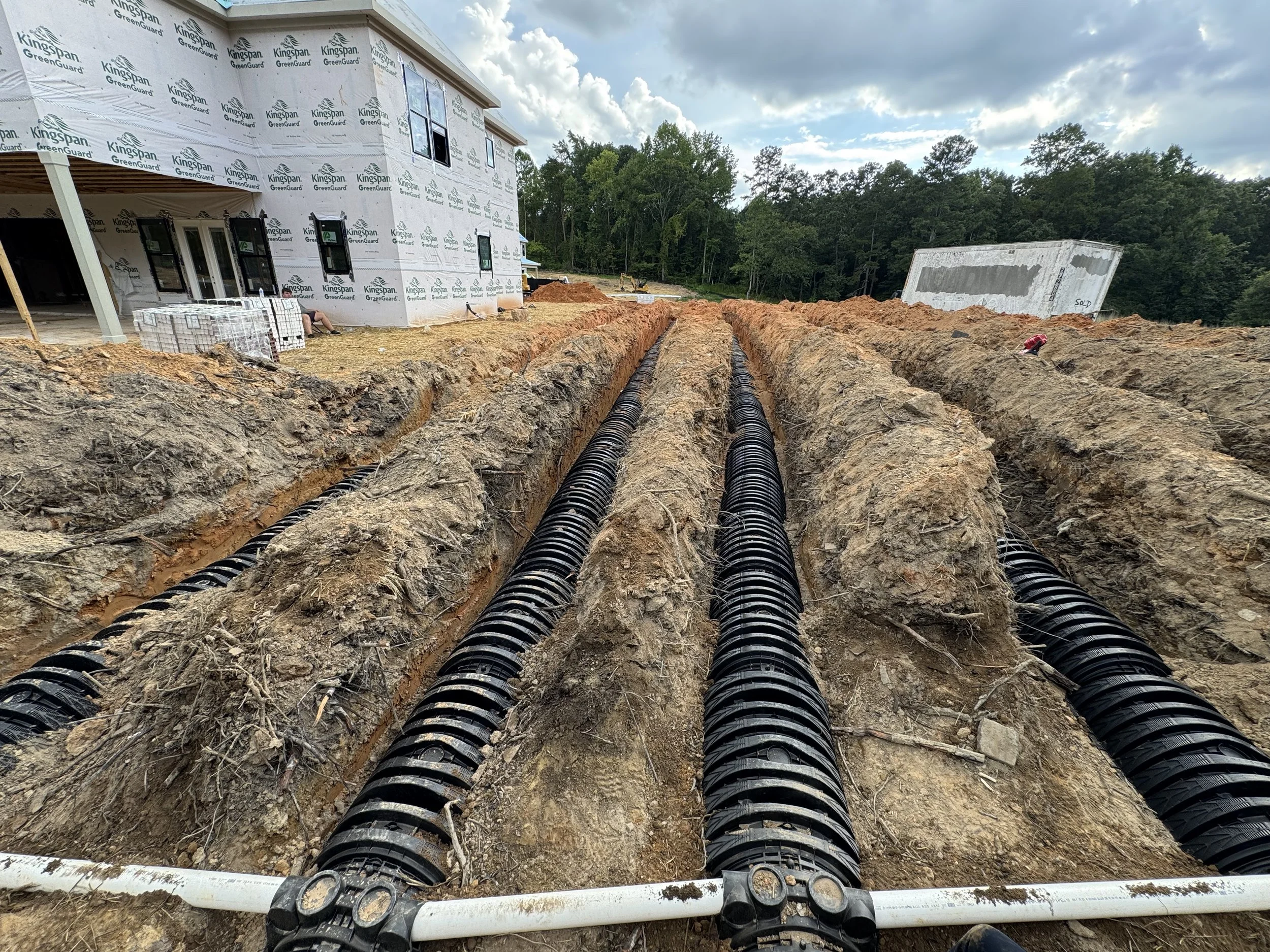 Underground piping installation at a construction site with a partially built house in the background and overcast sky.