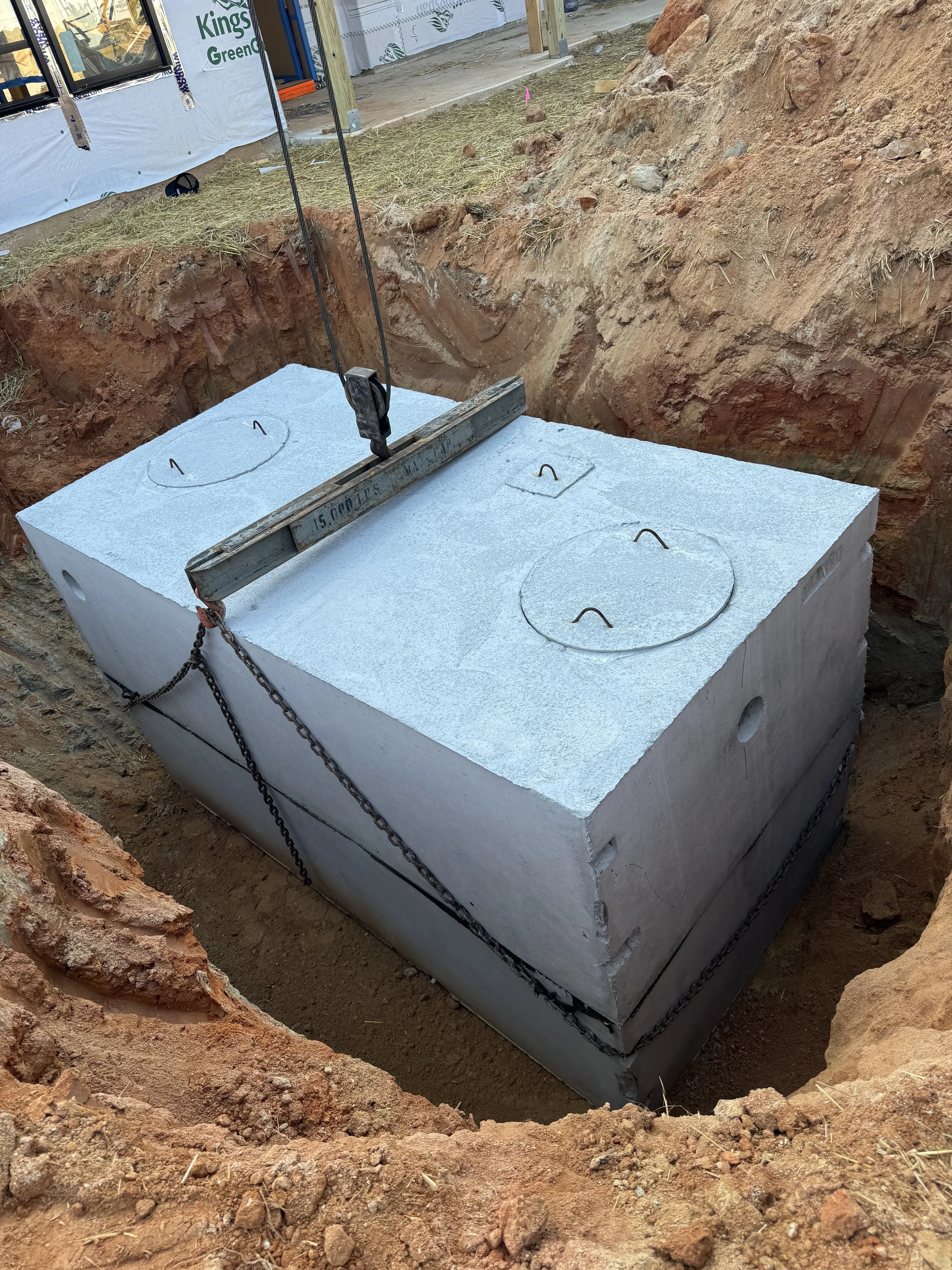 Large white concrete septic tank with circular cutouts on top, chains securing it in a dug-out hole, construction equipment and building under construction in the background.