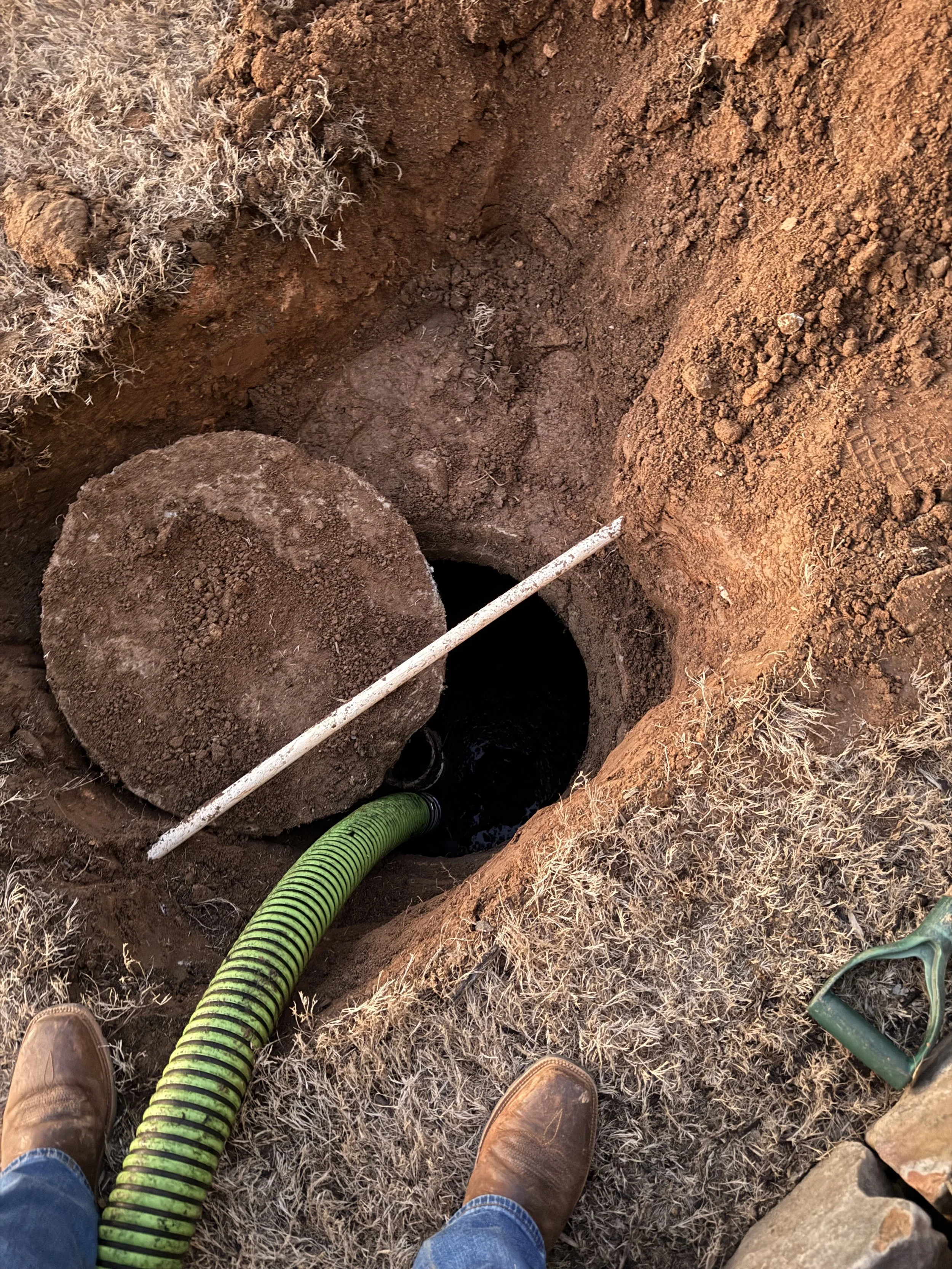 An open underground well with a large round cover removed, showing dark water inside. A green corrugated pipe is entering the well, and a long measuring stick is across the opening. The person's boots and jeans are visible at the edge of the trench, 