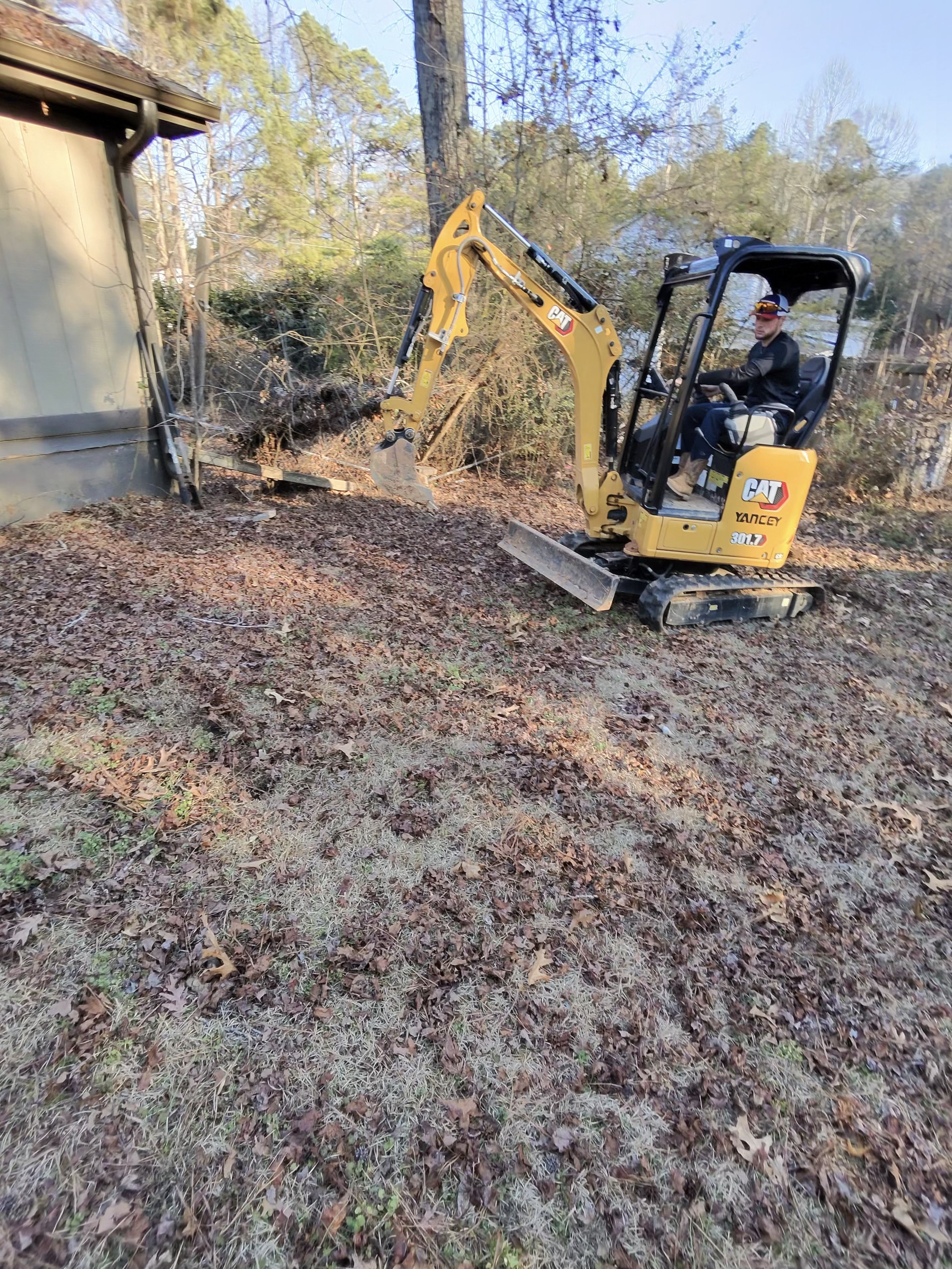 A person operating a yellow CAT mini excavator near a house, digging and clearing old leaves and debris from the ground in a wooded yard.