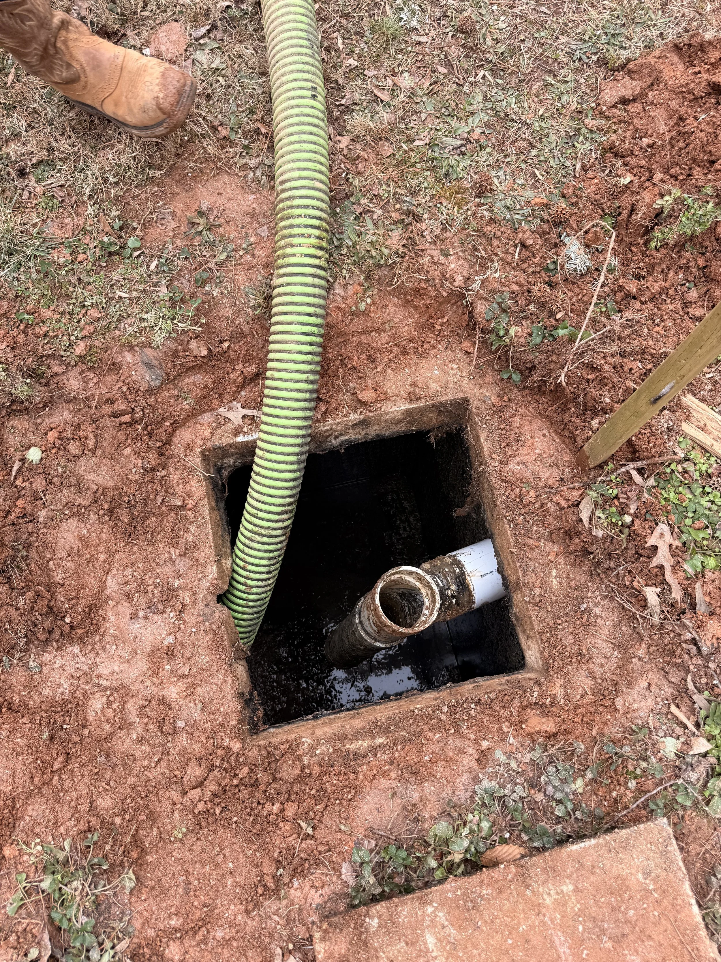 An outdoor view of a square hole in the ground with plumbing pipes inside, a green flexible drainage hose and a pipe with black and white sections, surrounded by red dirt and some small plants.