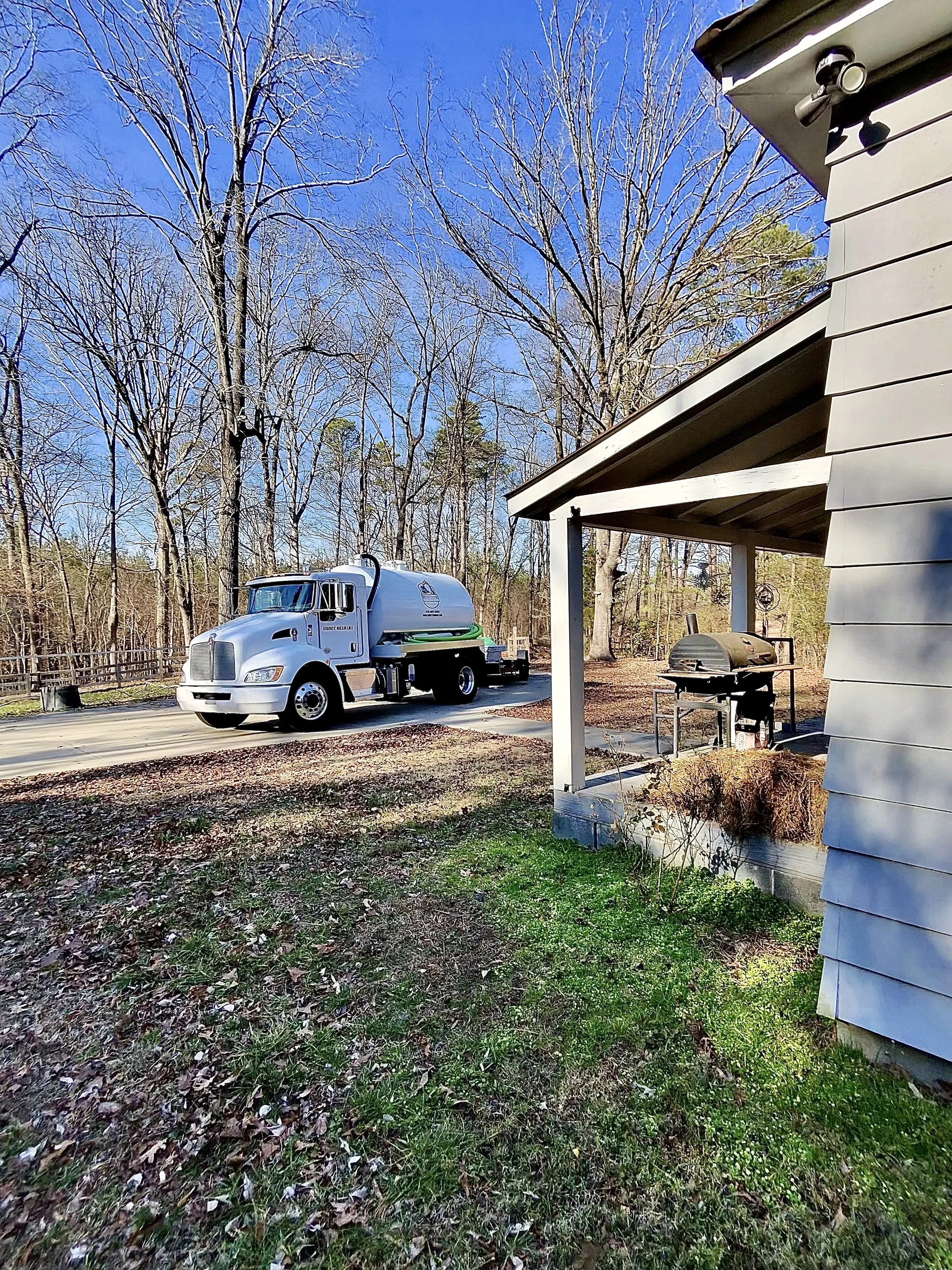 A white sanitation truck parked on a driveway with leafless trees and a partly cloudy blue sky in the background, next to a house with a porch and a charcoal grill.