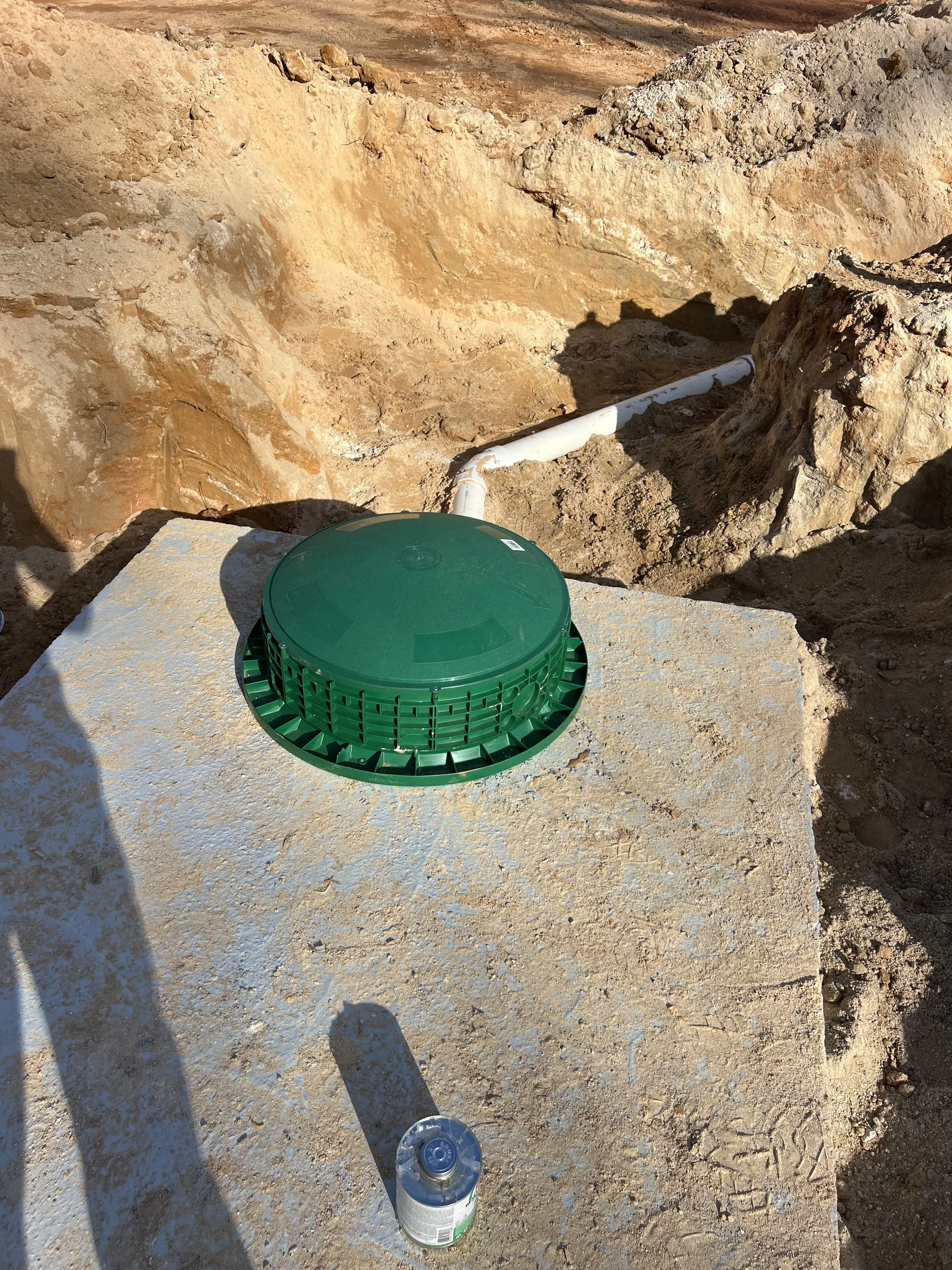 A septic tank platform with a green access lid, pipe connections, and a soda bottle on top of the platform, in a dirt construction site.