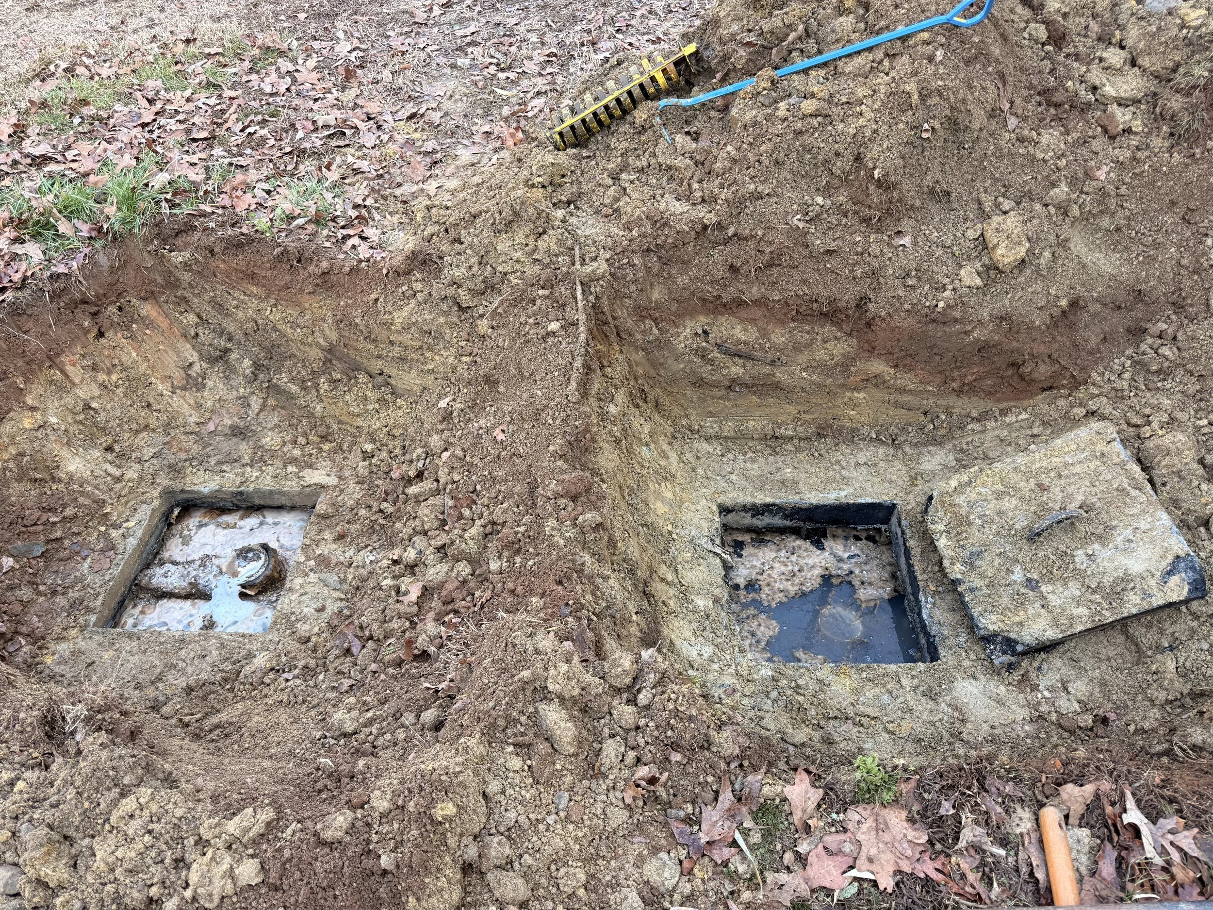 Two underground water meter boxes with covers, one open and one closed, surrounded by dirt and fallen leaves, with a yellow rake and a blue tool nearby.