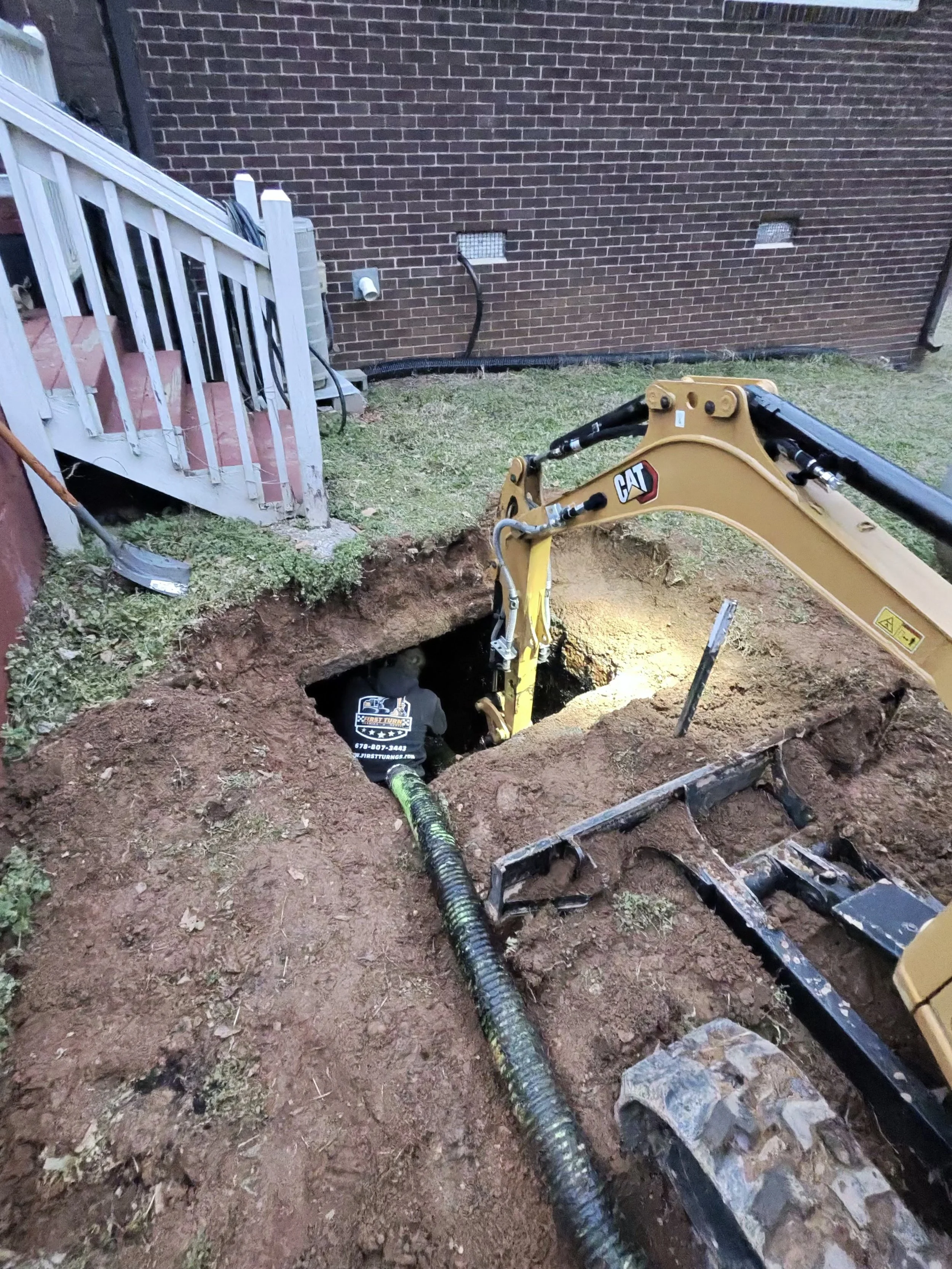 A construction worker operating a small excavator digging a hole next to a house with a brick wall, a small staircase leading to a door, and a black drainage pipe.