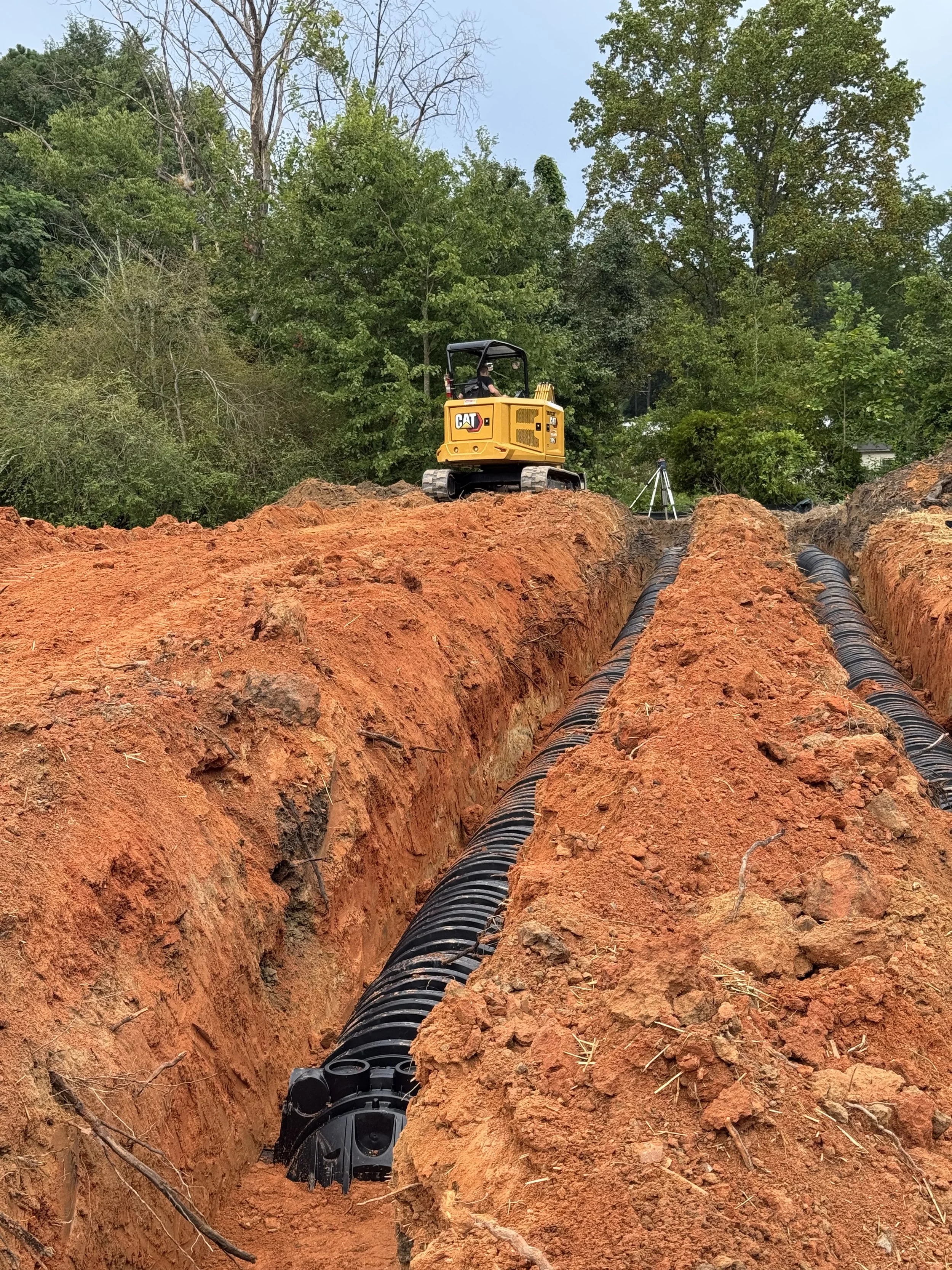 Construction site with a small yellow CAT excavator working on installing septic chambers for a drain field in a trench, with green trees and a partly cloudy sky in the background.