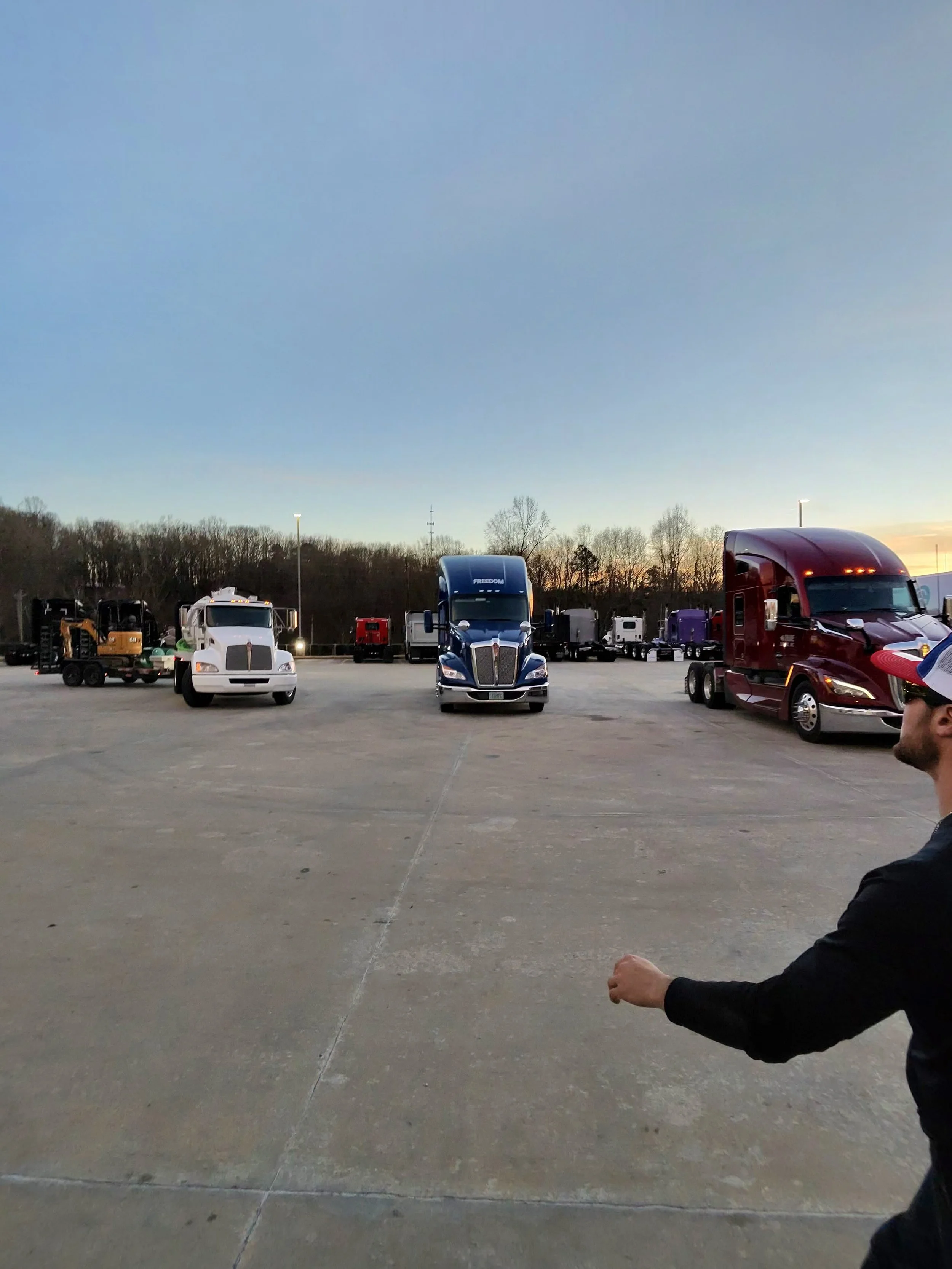 A lineup of semi-trucks in a parking lot during sunset with a man in a black jacket and cap visible on the right side.
