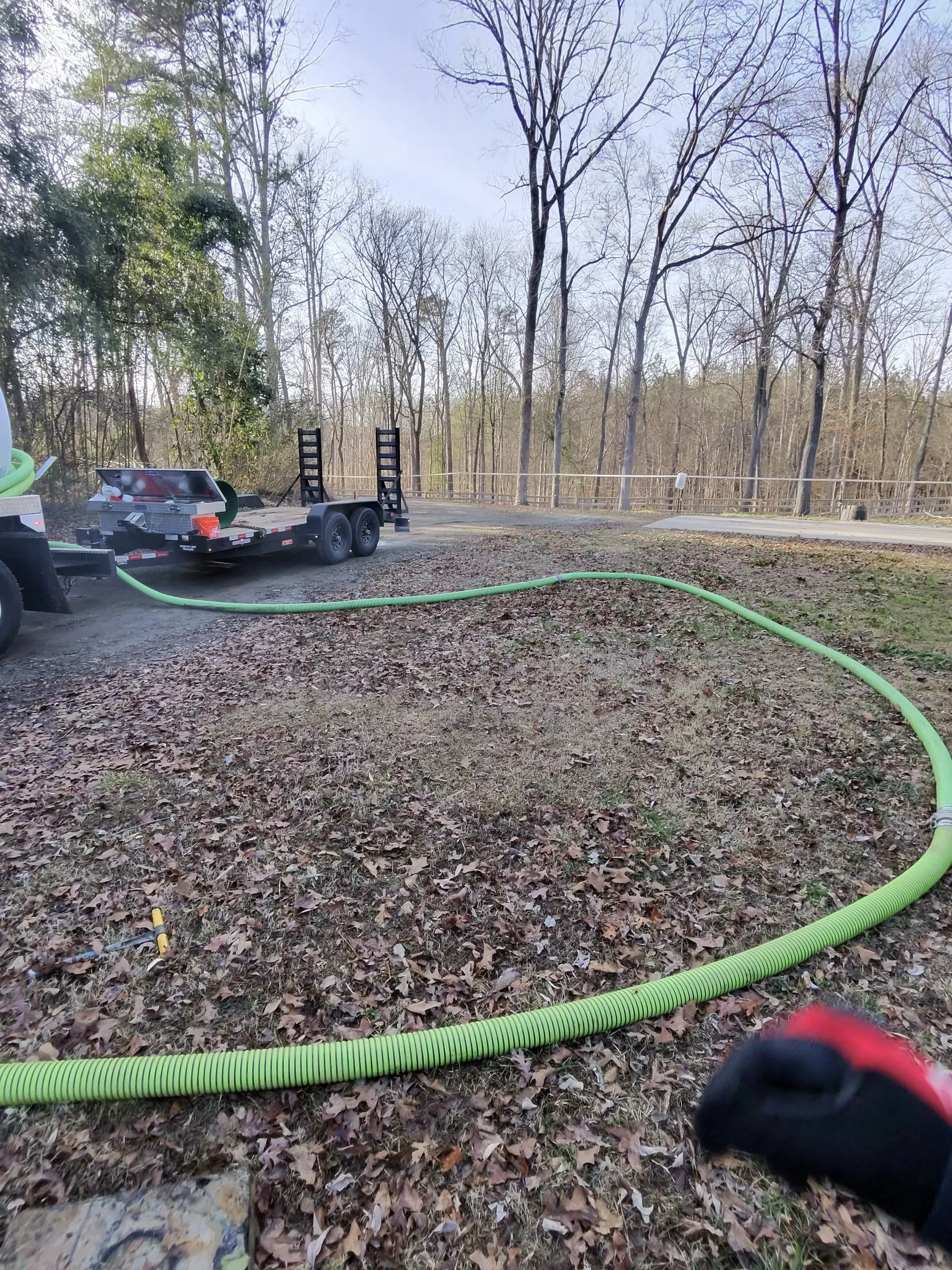 A green hose connected to a large tank on a trailer, lying on a dirt ground covered with fallen leaves, with leafless trees in the background and a paved road in the distance.