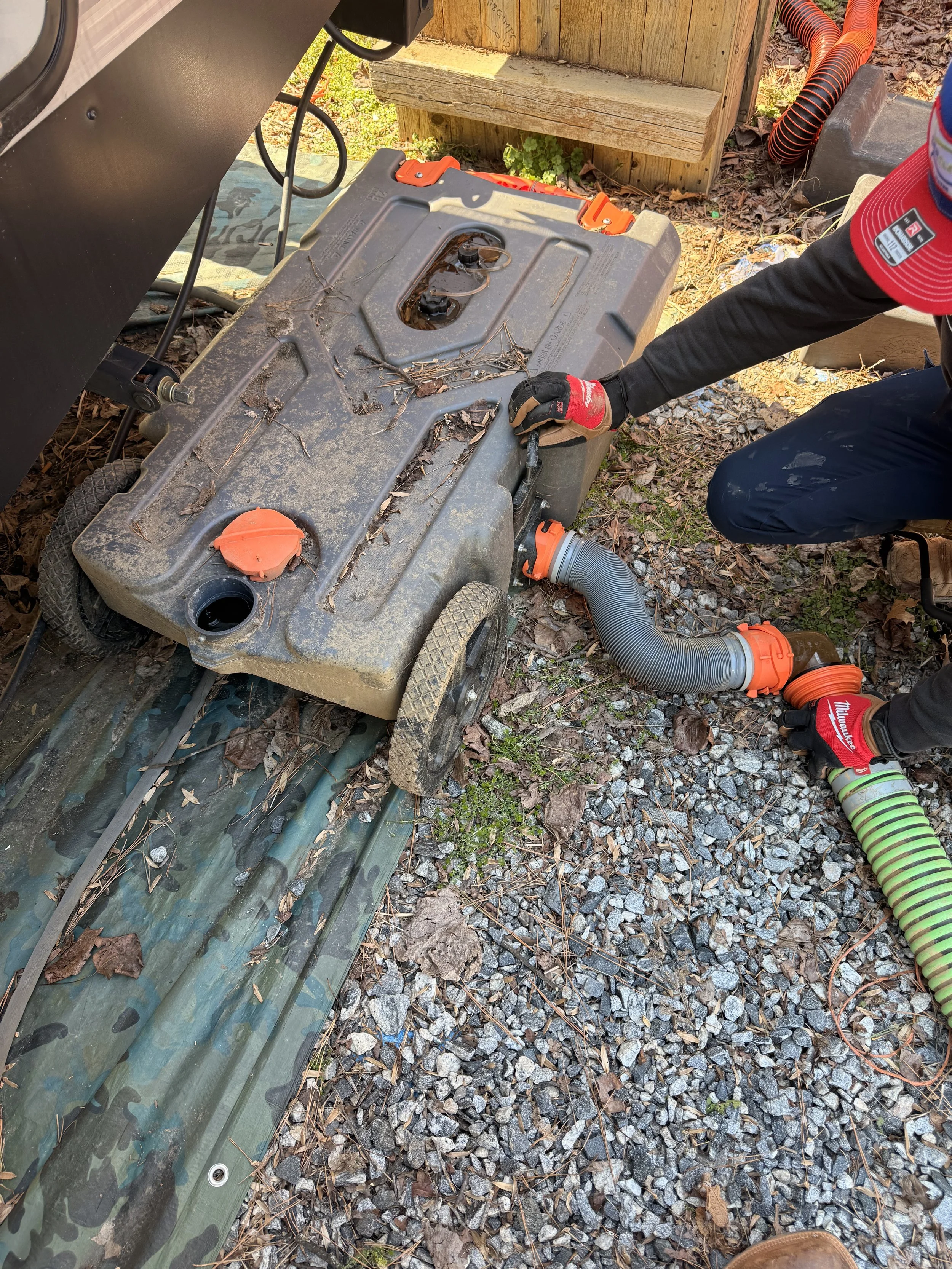 A person working outdoors on a septic tank or similar underground tank, connecting large flexible hoses for the system, with gravel and leaves on the ground.